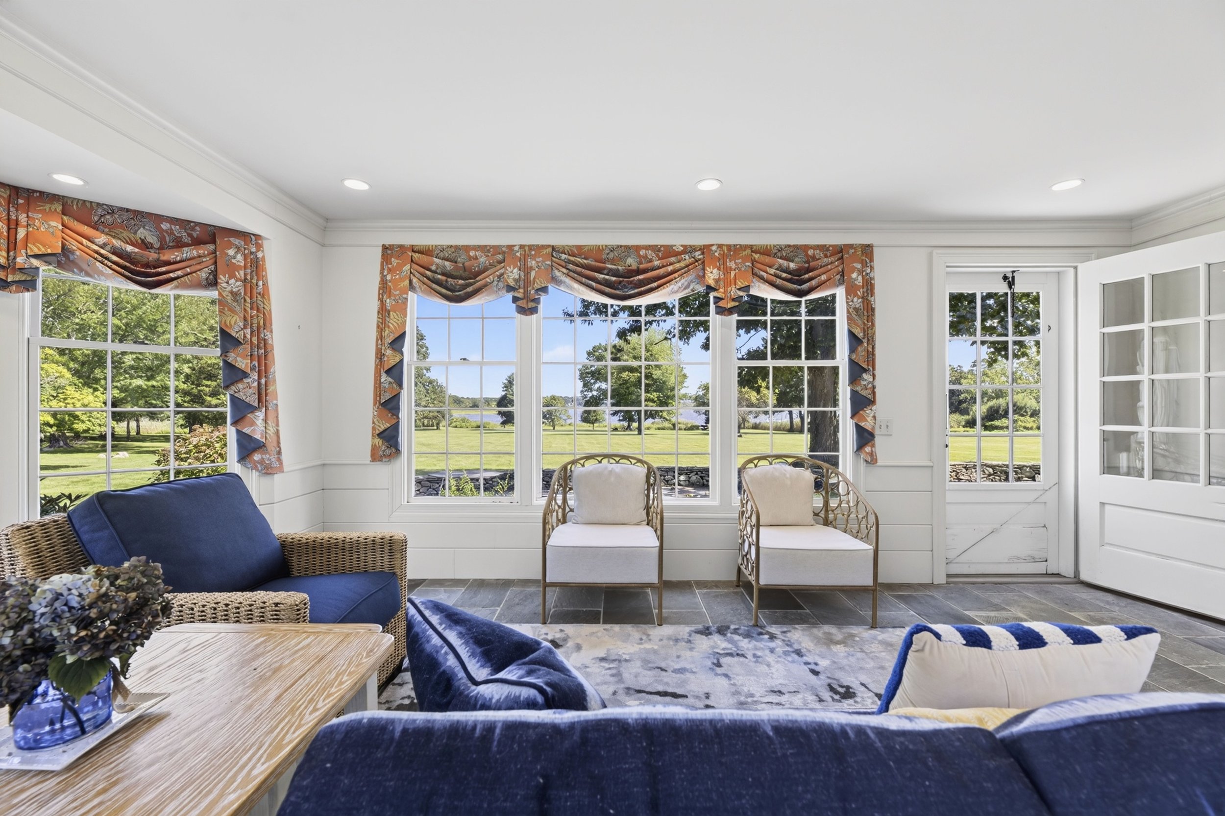 Bright living room with large windows, floral curtains, wicker chairs, a blue sofa, and a view of a green yard outside.