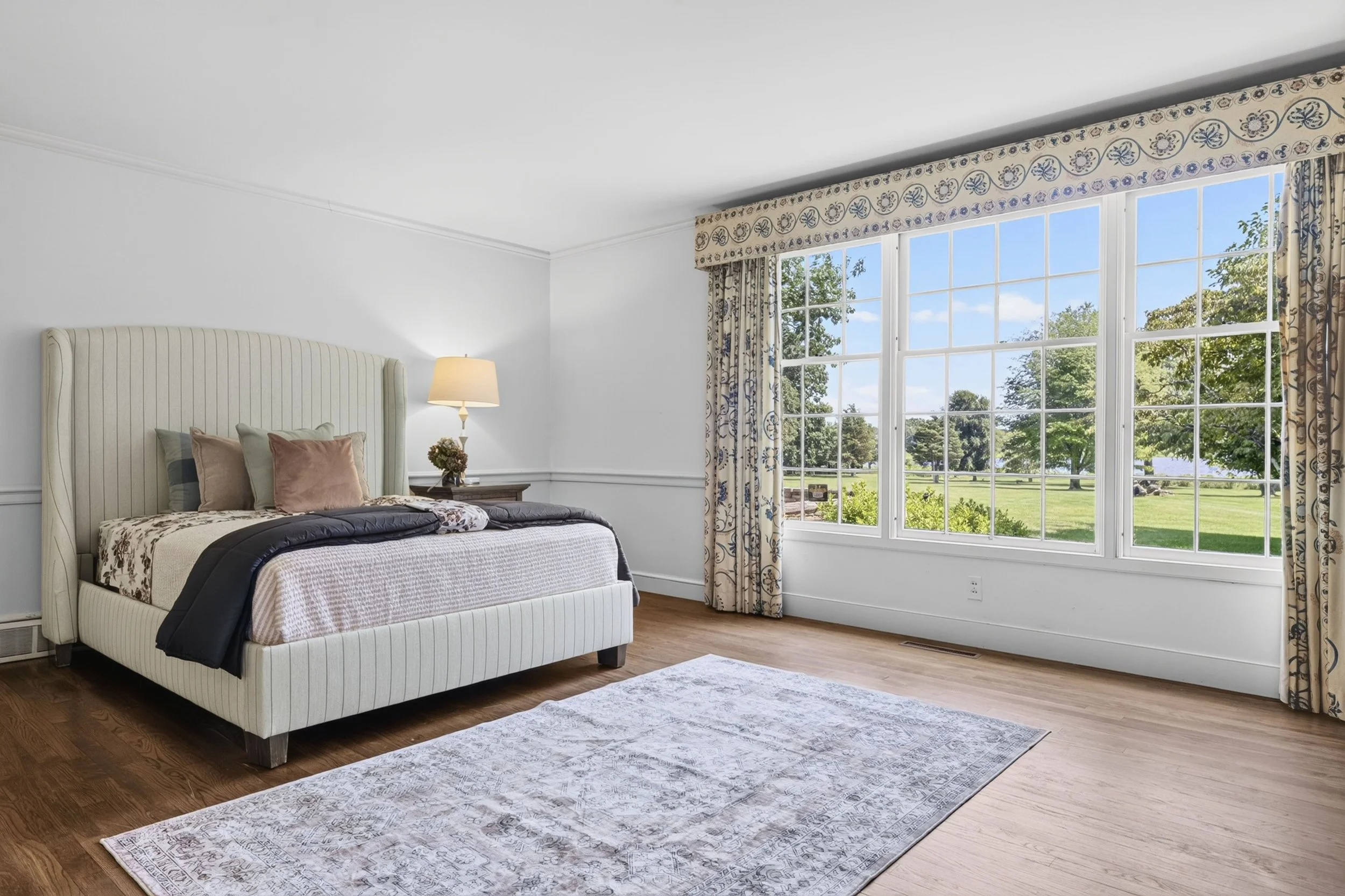A bedroom with a beige upholstered bed, neutral pillows, a nightstand with a lamp, a window with patterned curtains, a view of green trees and a lake outside, and a light-colored rug on wooden flooring.