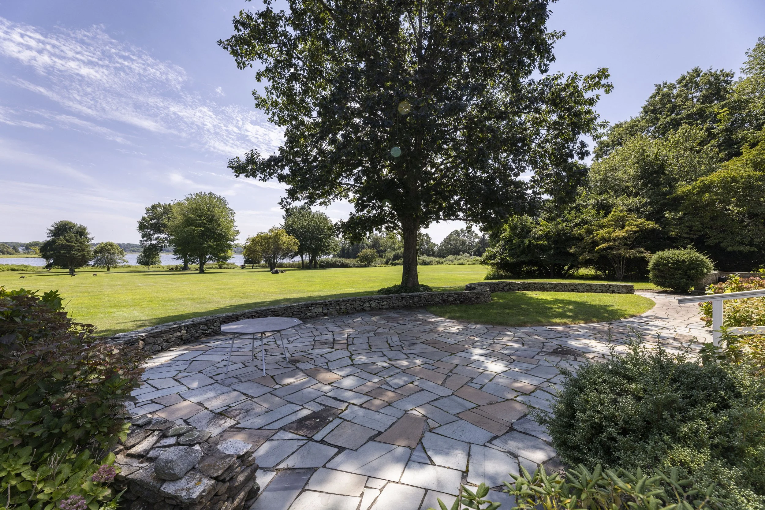 A scenic view of a backyard with a curved stone patio, lush green grass, and tall trees under a blue sky with wispy white clouds.