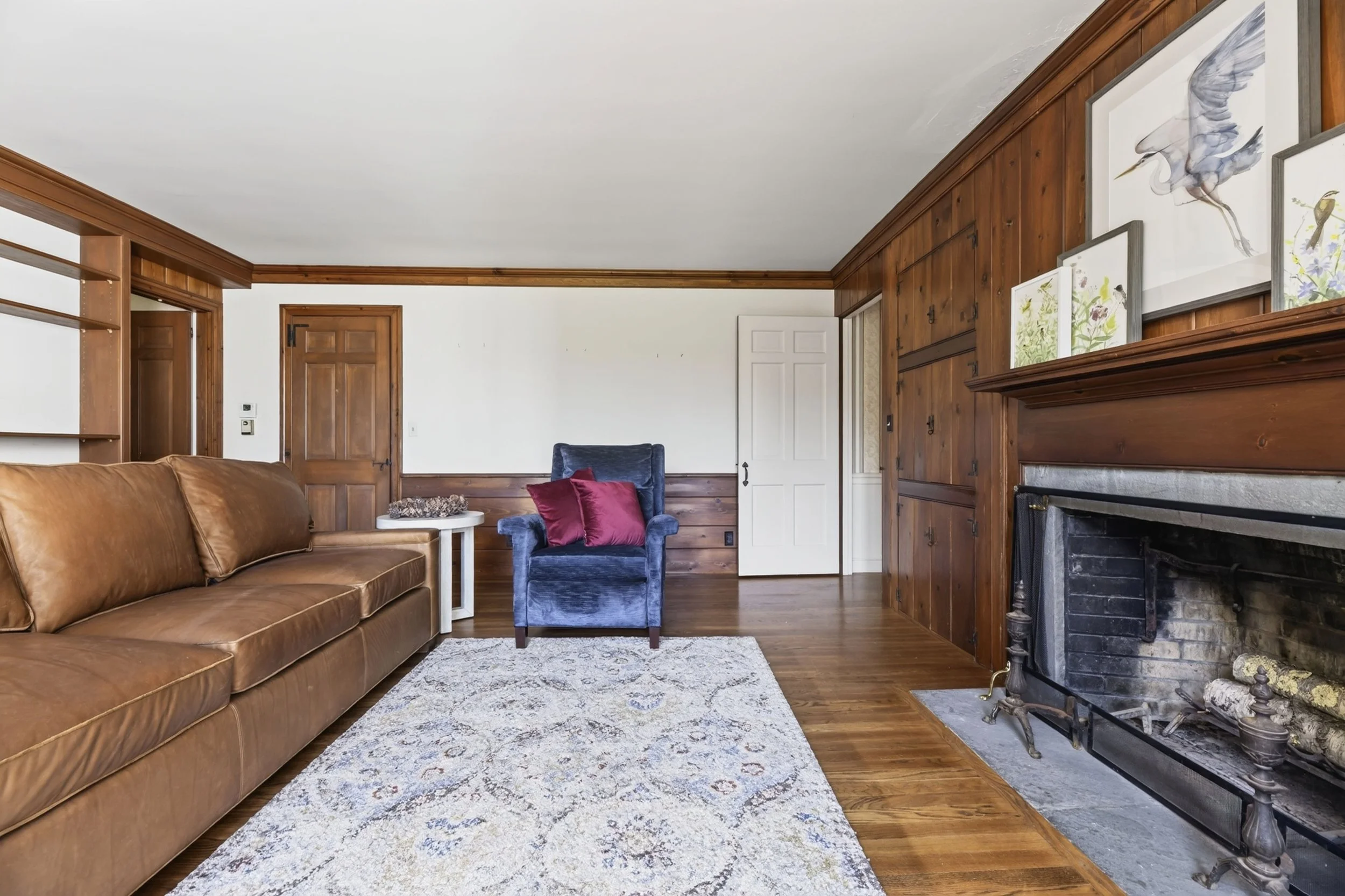 A cozy living room with a leather sofa, a blue velvet armchair with red pillows, a white side table, a multicolored patterned rug, a wood-paneled wall with framed artwork of birds, a white door, and a fireplace with logs.