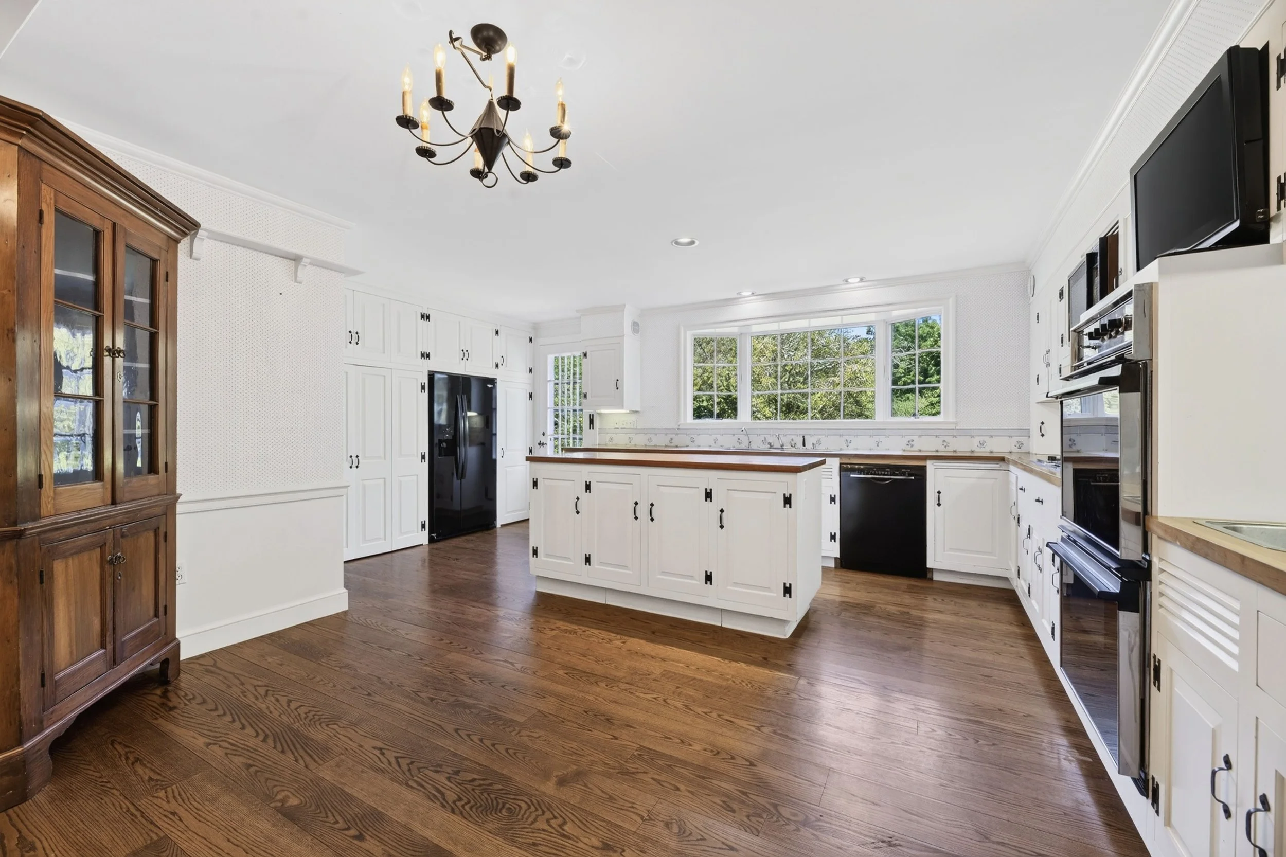  photographed view of a bright kitchen with white cabinets, wooden countertop, large windows, hardwood floors, and a ceiling chandelier.
