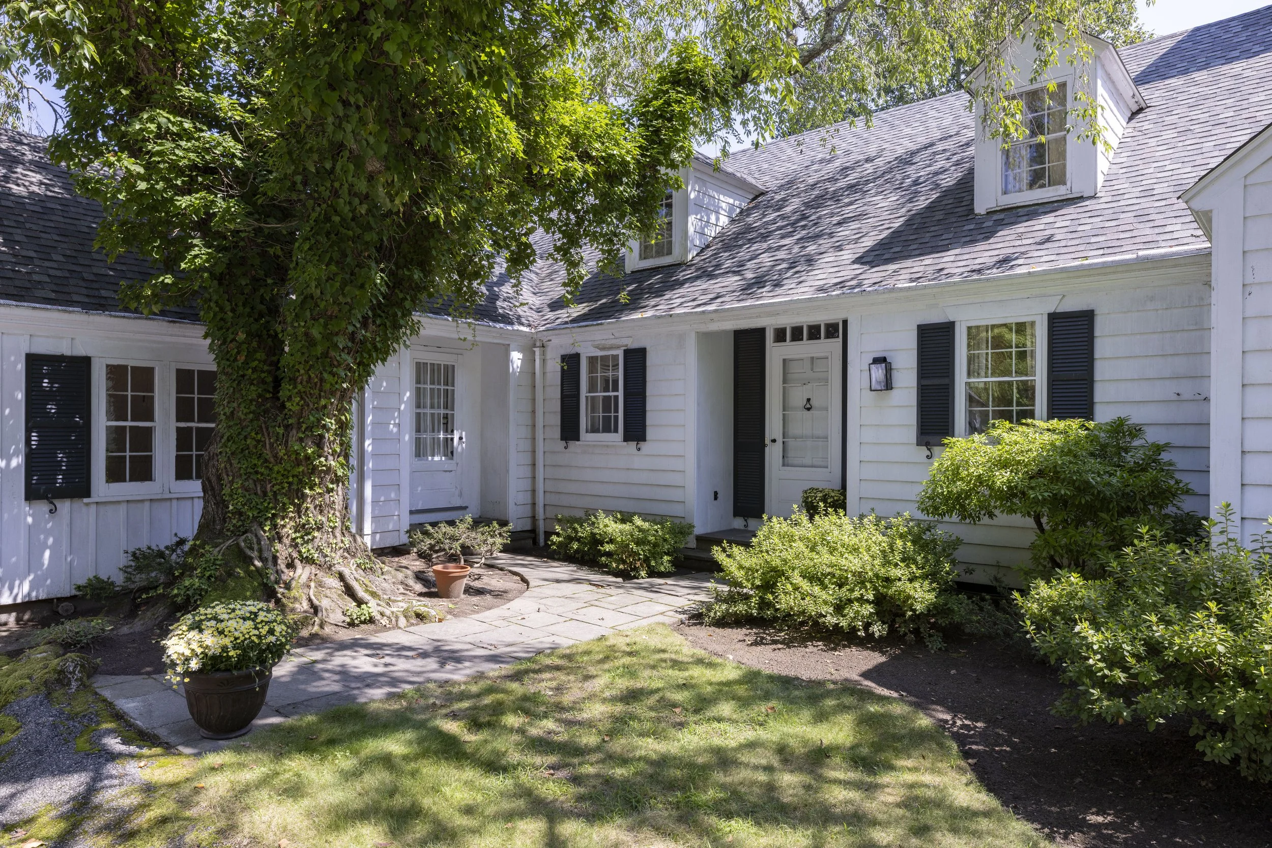 White house with black shutters and dormer windows, surrounded by greenery and a large tree in the front yard.
