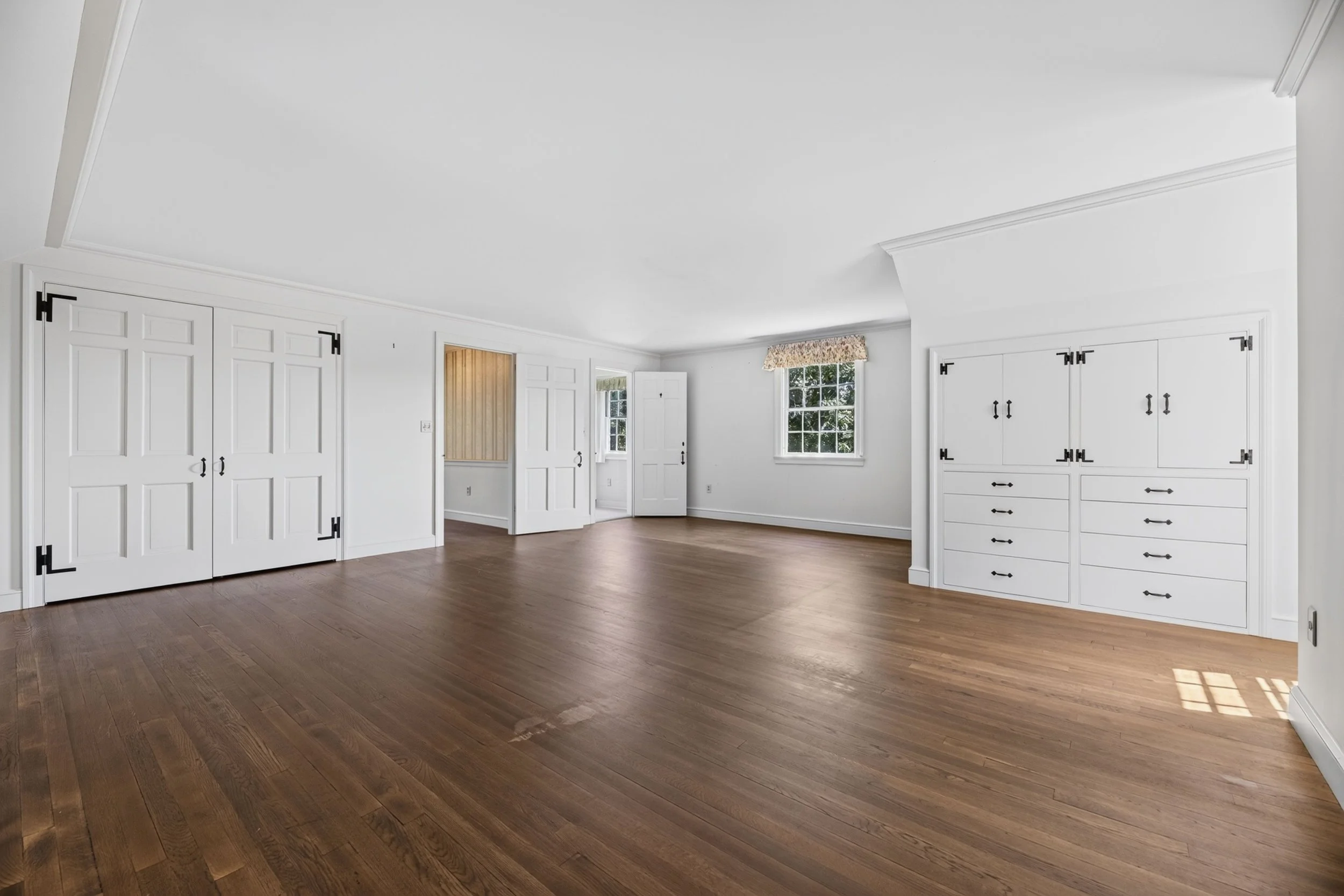 Empty room with white walls, wooden floors, multiple built-in white cabinets, a window with a floral valance, and a partly open door.