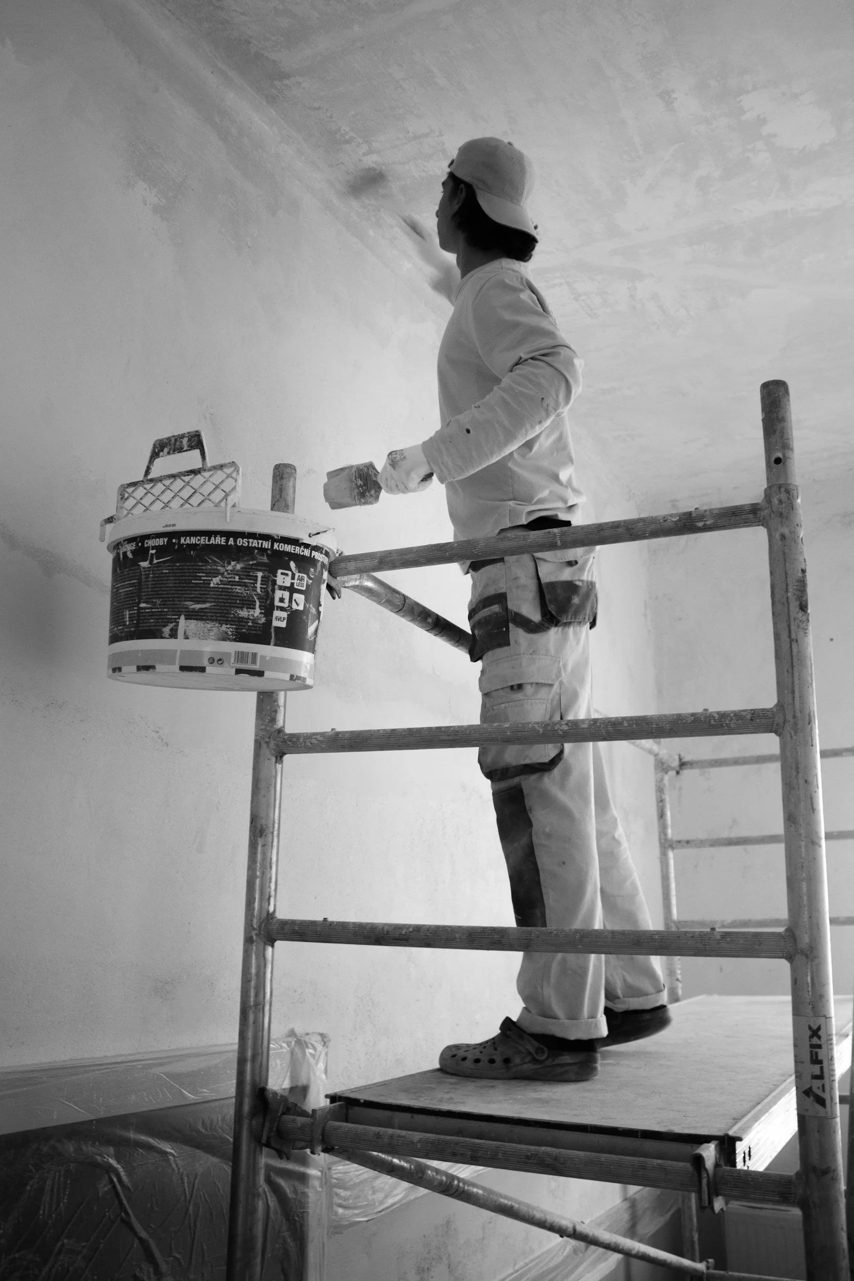 A person wearing a hat and work clothes stands on scaffolding, painting or repairing a ceiling with a roller. There is a bucket of paint or plaster nearby.