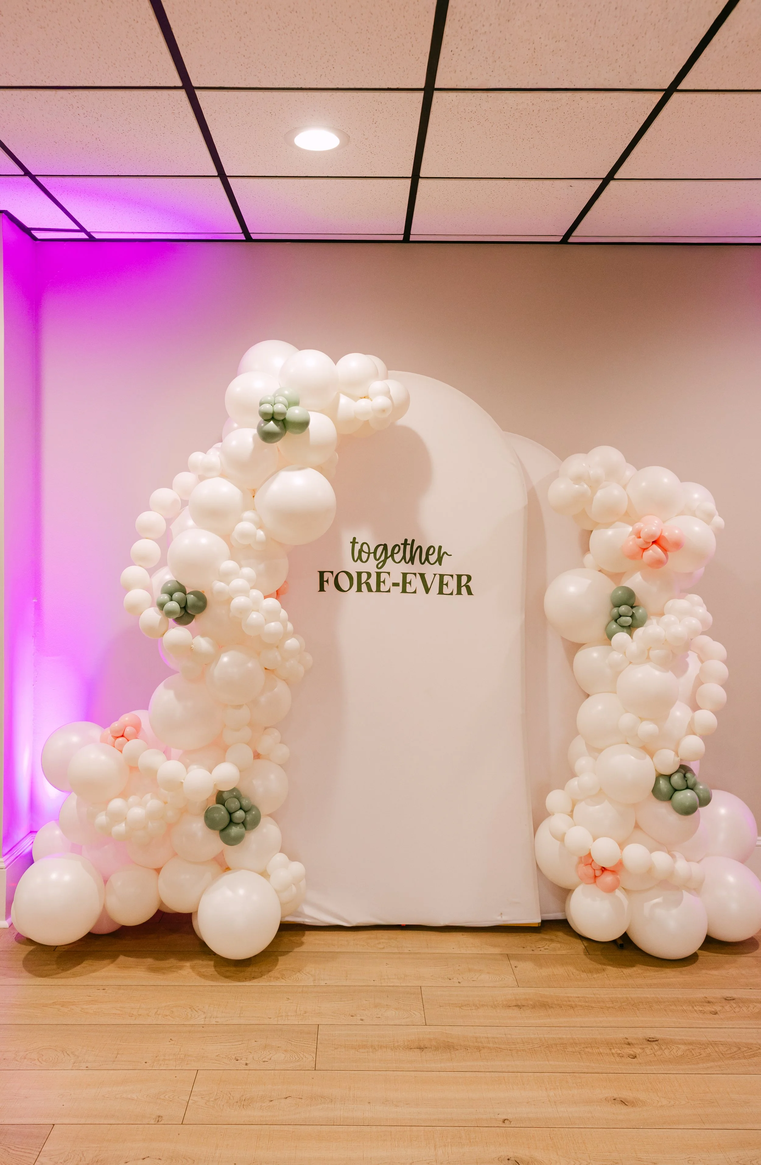 A balloon decoration with white, pink, and green balloons arranged around a backdrop that says 'together FORE-VER' in black and green text, with purple lighting on the left side.