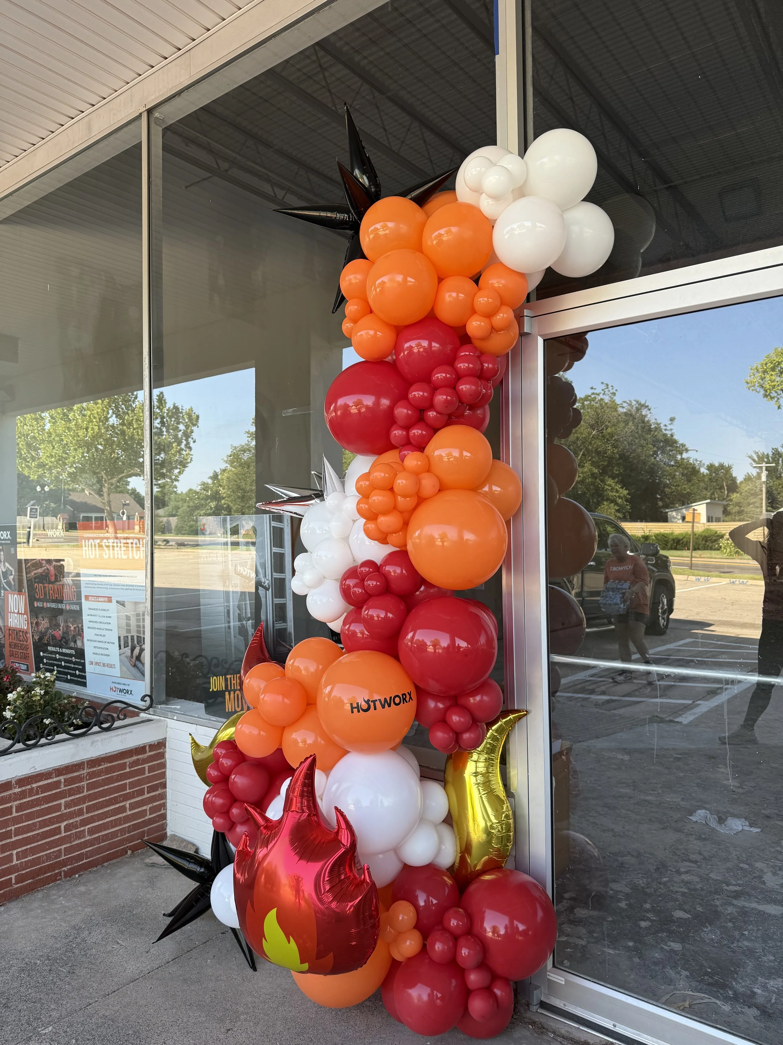 Decorative balloon arrangement with red, orange, black, and white balloons on the side of a building. The balloons form a cascading column and include some star-shaped and moon-shaped balloons. The building has large glass windows and a brick base, w