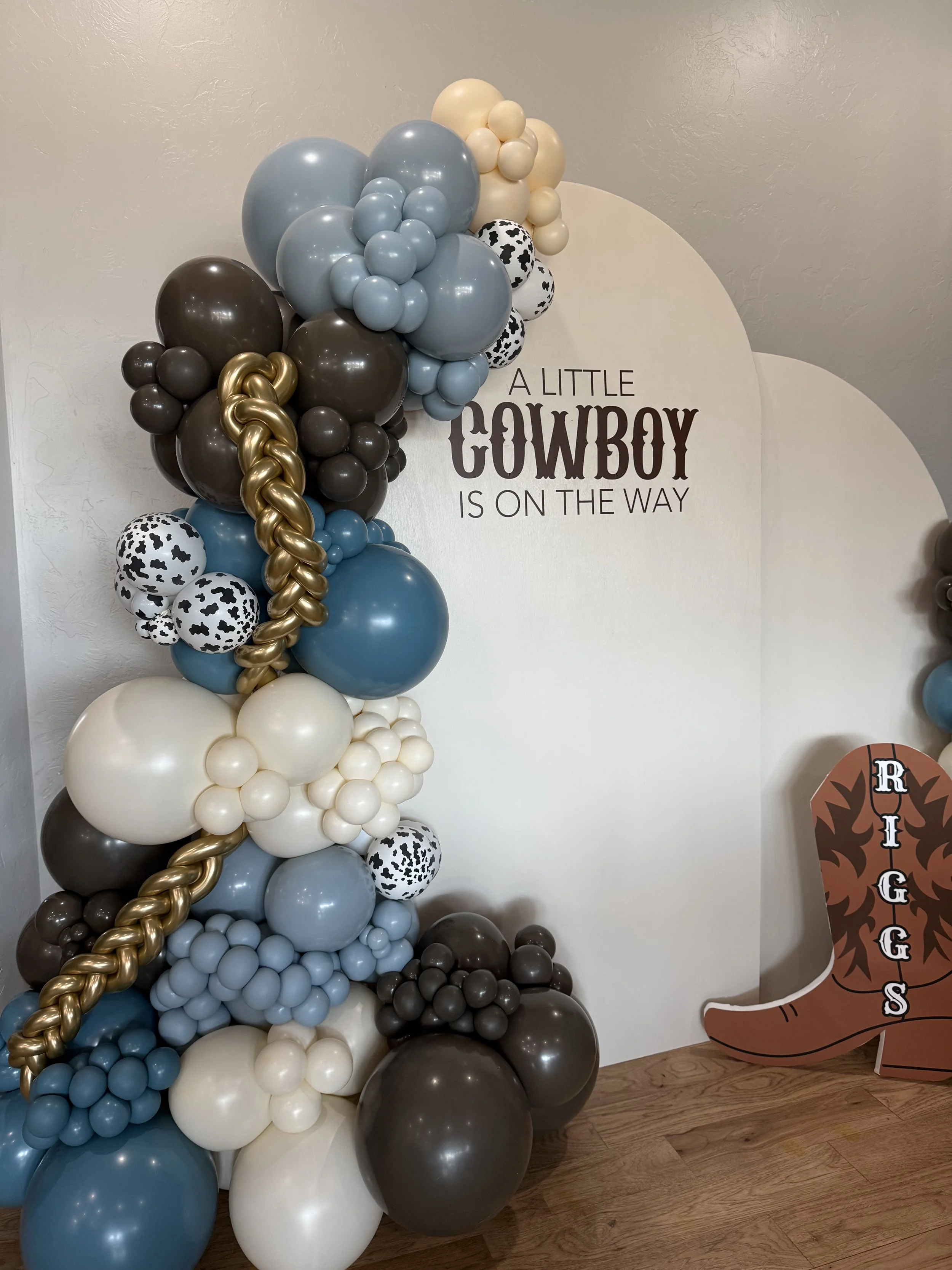 A balloon arrangement with various blue, white, brown, black, and gold balloons forming an arch or decorative display next to a sign that reads "A Little Cowboy Is On The Way." There is also a cardboard cowboy boot cutout decorated with silver, black