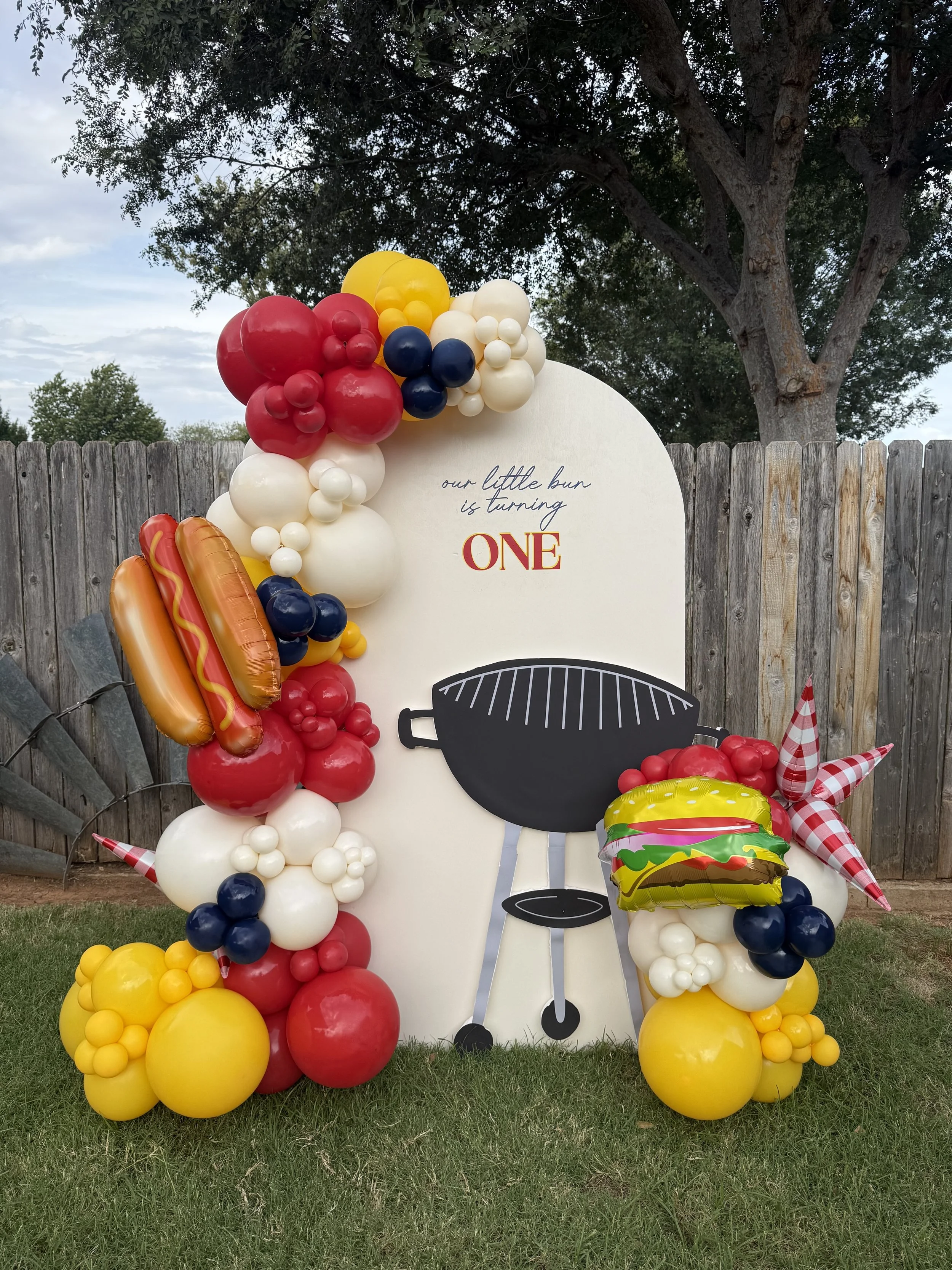 Decorative birthday display with colorful balloons arranged around a white arched board that has the message "our little bun is turning ONE." The display includes balloon hot dogs, burger, star-shaped balloon, and a black grill cutout, set outdoors against a wooden fence and a tree.