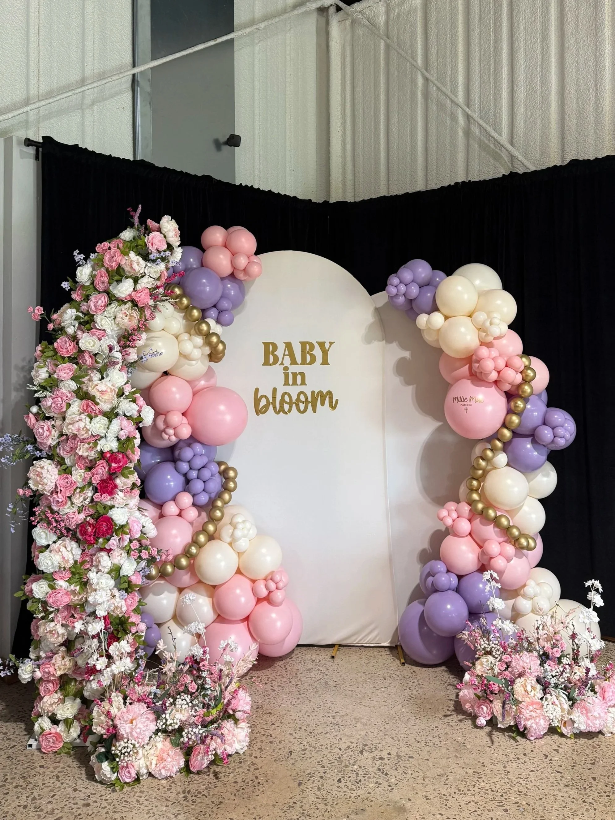 Decorative floral display with pink, purple, white balloons and flowers, with a sign reading 'Baby in Bloom' at a celebration or event.