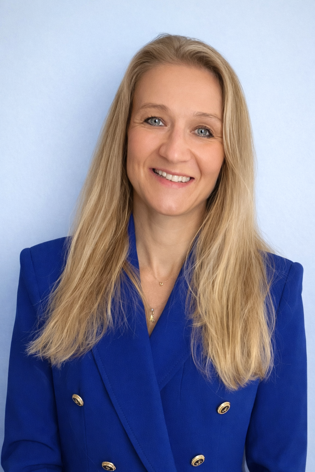 A woman with long blonde hair wearing a blue blazer and smiling, standing in front of a light blue background.