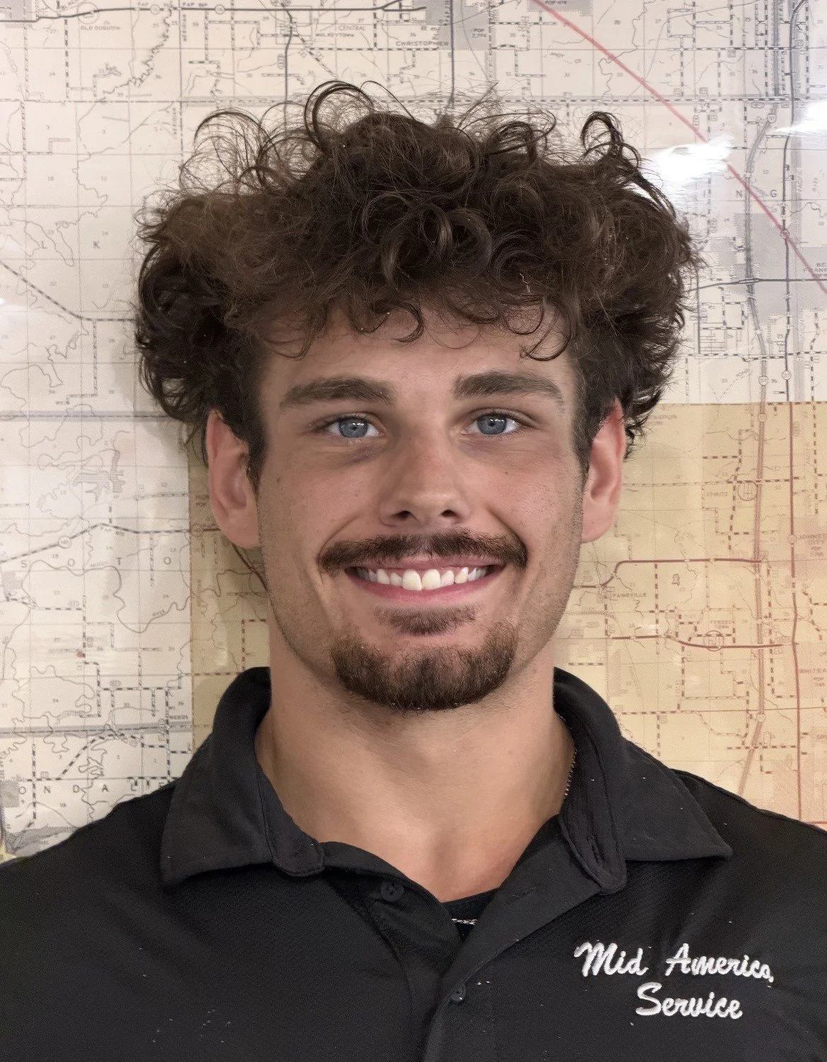 A smiling young man with curly hair and a mustache, wearing a black shirt with white embroidery that reads 'Mid-America Service,' standing in front of a wall with a detailed map.