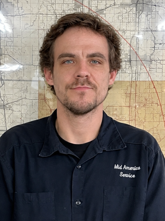 A man with curly brown hair and blue eyes gazing at the camera, wearing a navy blue shirt with "Mid America Service" embroidered on it, standing in front of a map background.