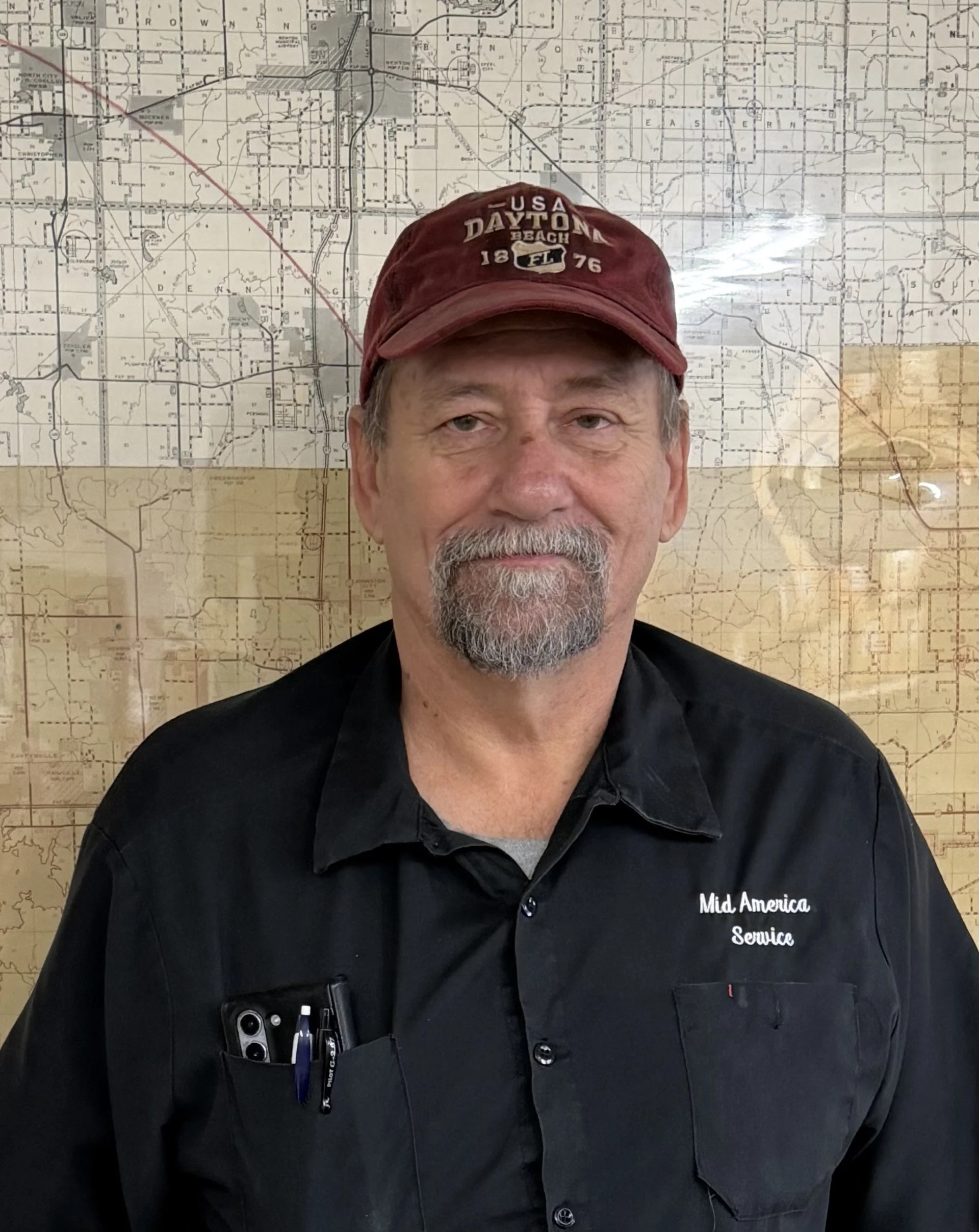 A man with a beard and mustache wearing a red cap that says 'USA Daytona Beach 1876 Florida' and a black shirt that says 'Mid America Service' in front of a large map or blueprint on the wall.