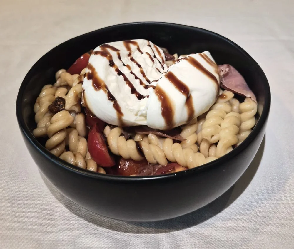A bowl of pasta topped with burrata cheese, drizzled with balsamic glaze, and possibly some cured meats with cherry tomatoes.