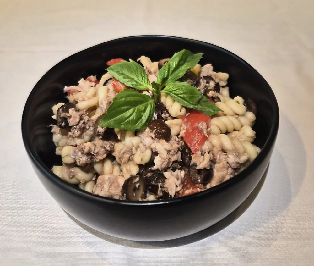 Black bowl of pasta salad with black olives, ground meat, tomatoes, and fresh basil leaves on top.