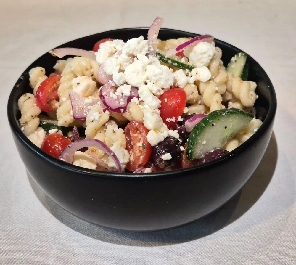 A bowl of pasta salad with cherry tomatoes, cucumbers, red onions, black olives, feta cheese, and pasta, in a black bowl on a light surface.