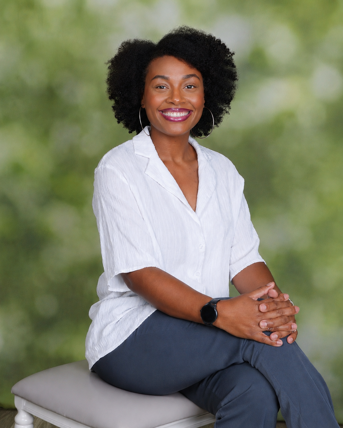 A young woman with curly black hair, smiling, wearing a white button-up shirt, dark pants, hoop earrings, a wristwatch, and sitting on a light-colored stool with a green blurred background.