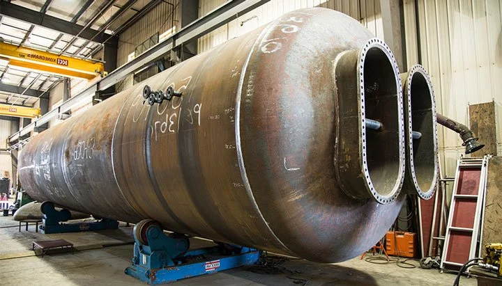 Large steel pressure vessel with two flanged nozzle openings, on rotating stands in the Penfabco fabrication shop