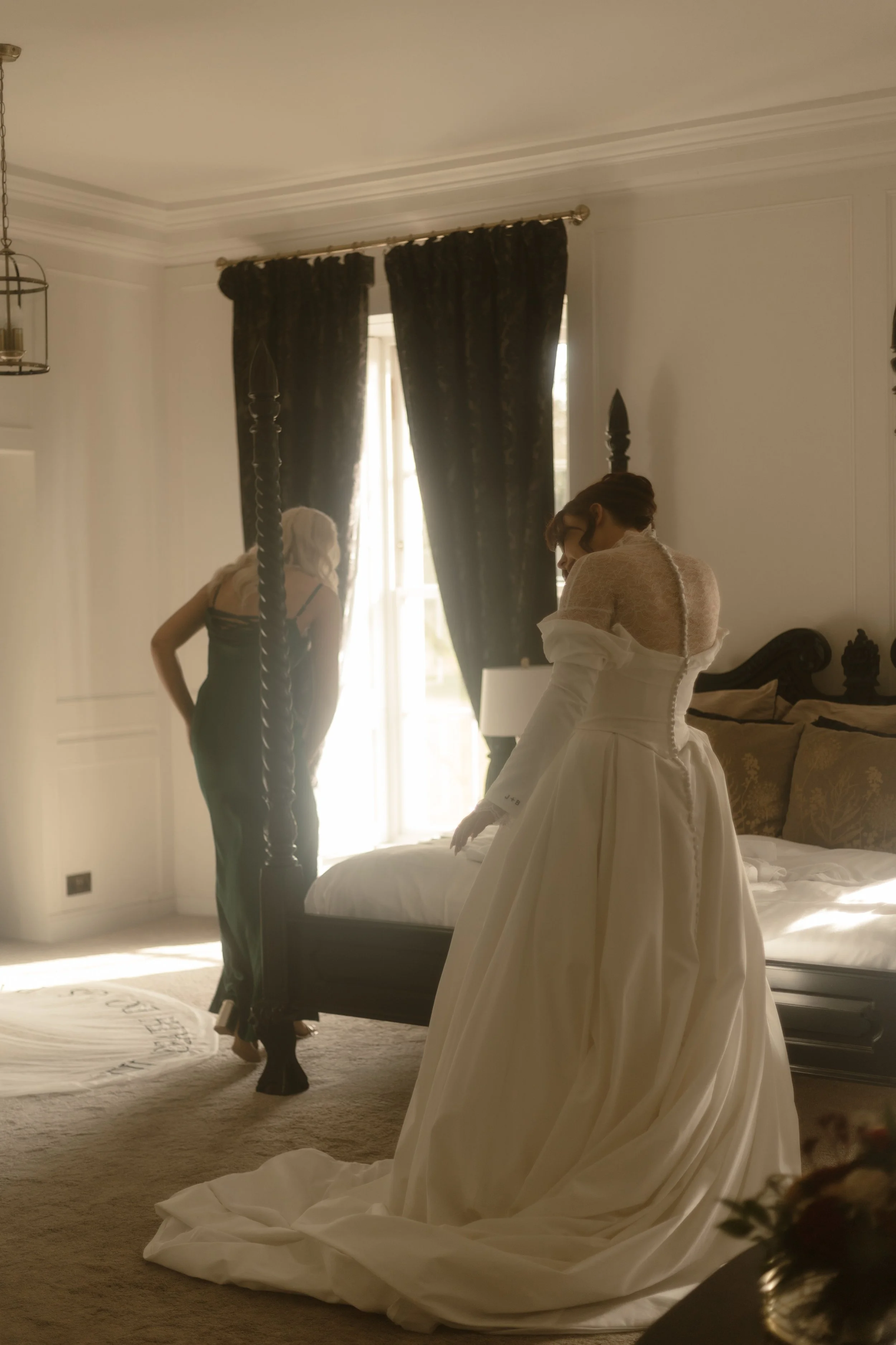 A bride in a white wedding dress sitting on a bed, with her back to the camera, in a room with dark curtains and a woman in black attire standing near a window.