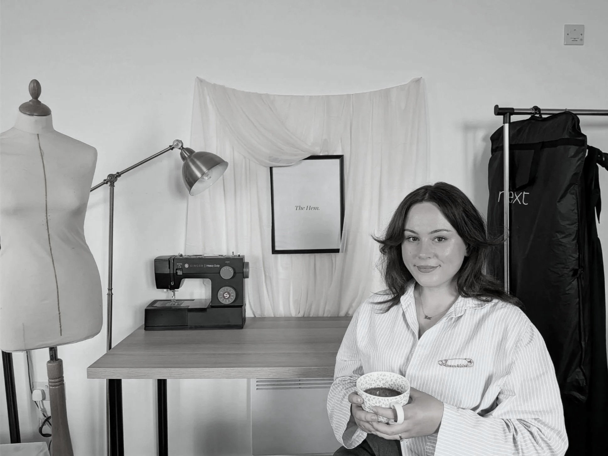 A woman with brown hair and a striped shirt holds a bowl with both hands, standing in a sewing or craft room. The room features a mannequin, a sewing machine, a framed quote "The Hem", a lampshade, and a clothing rack with black bags.