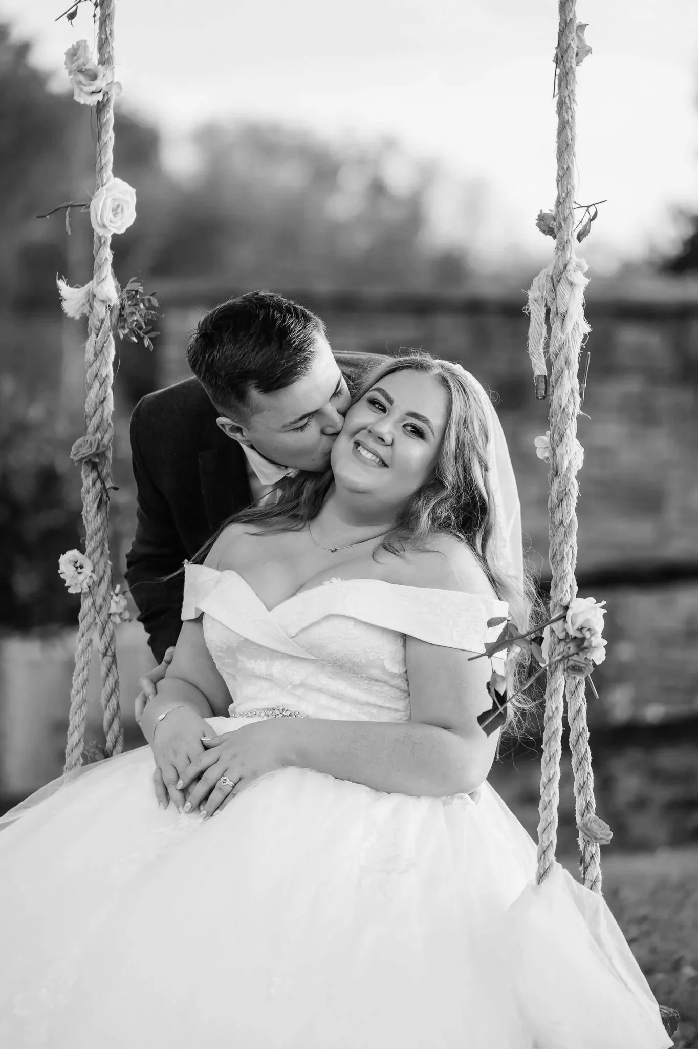 A black and white photograph of a couple on a swing. The woman is seated, smiling with her hair down, wearing a strapless wedding dress and an engagement ring. The man is leaning in, kissing her cheek, dressed in a suit. The swing is decorated with flowers and hanging ropes, with an outdoor natural background.