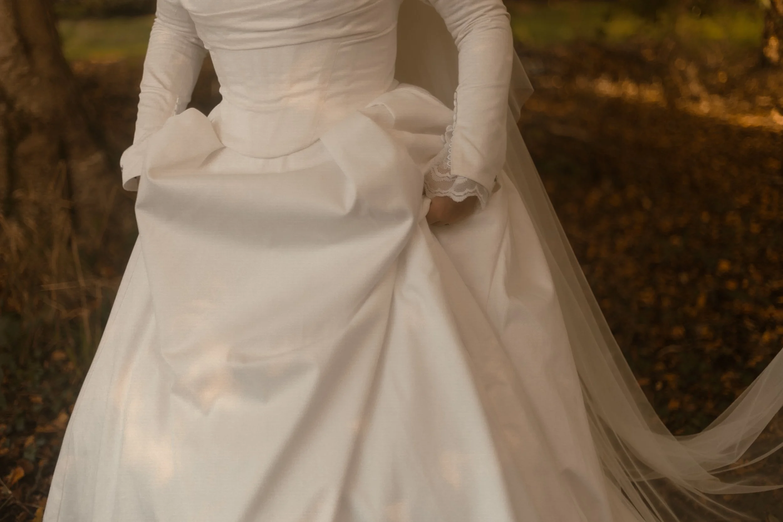 Close-up of a bride in a white wedding dress with lace accents, standing outdoors in a wooded area with fallen leaves.