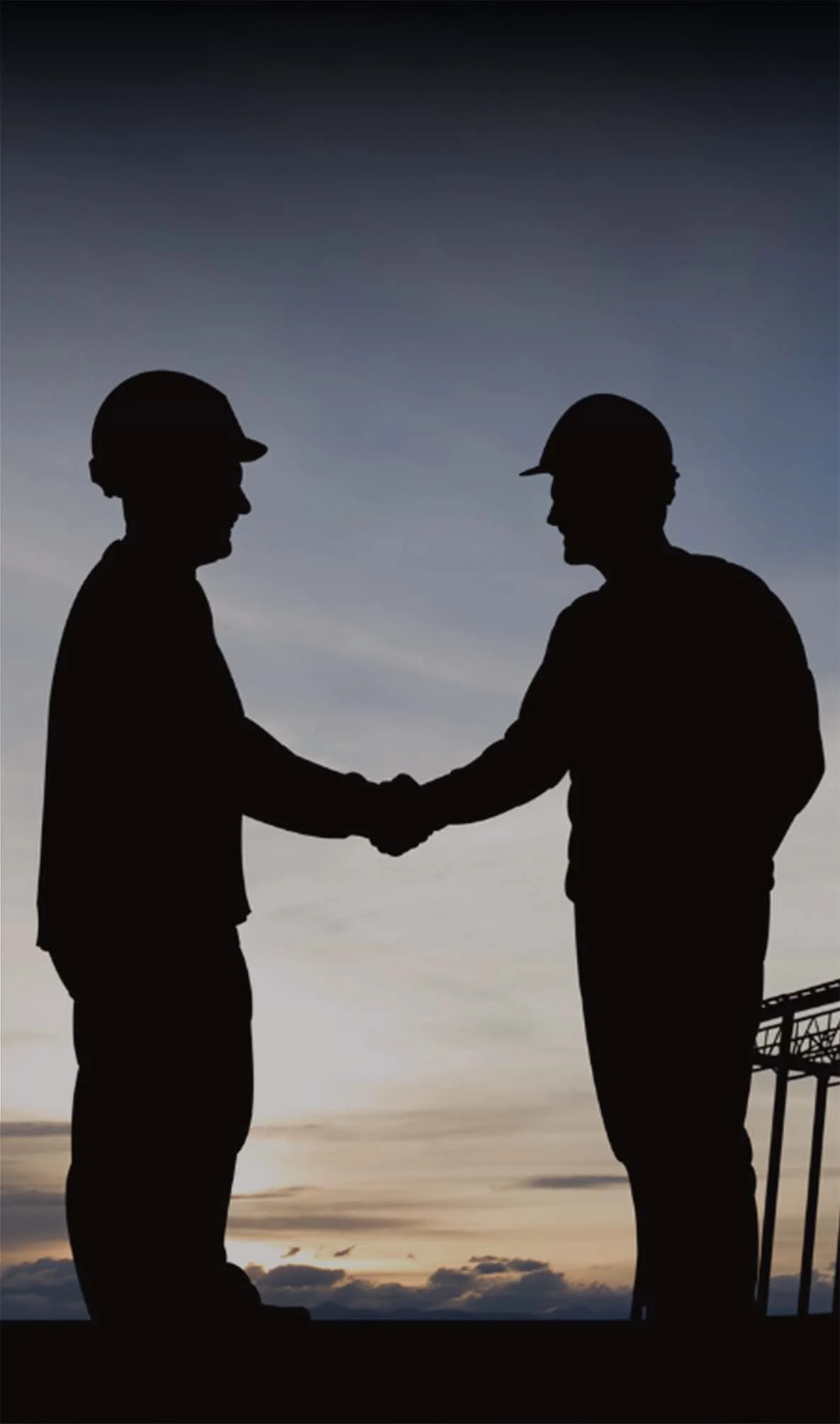 Silhouette of two men in hard hats shaking hands outdoors during sunset, with a clear sky and a cityscape and street lamp in the background.