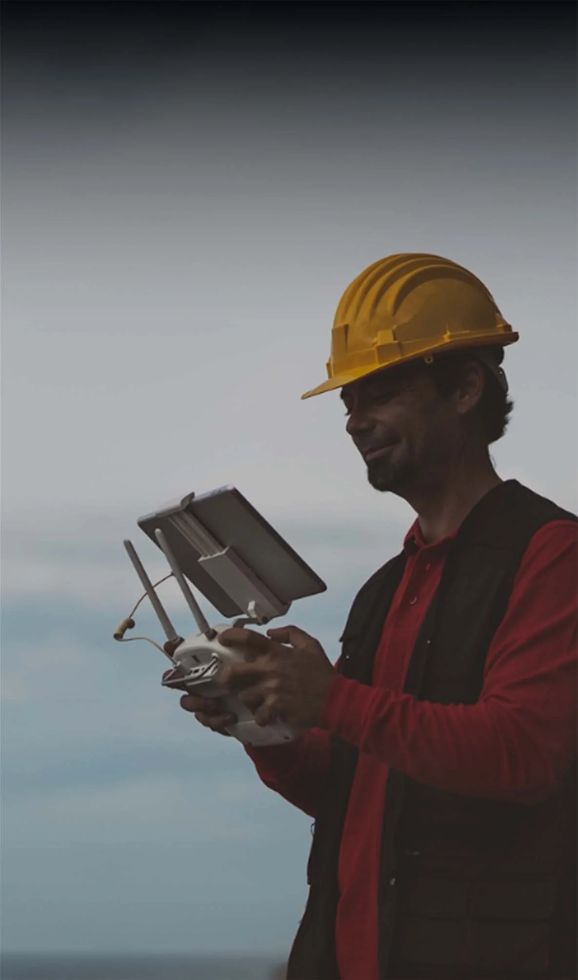 A man wearing a yellow safety helmet and red and black clothing is holding a remote control drone with a screen, against a cloudy sky background.