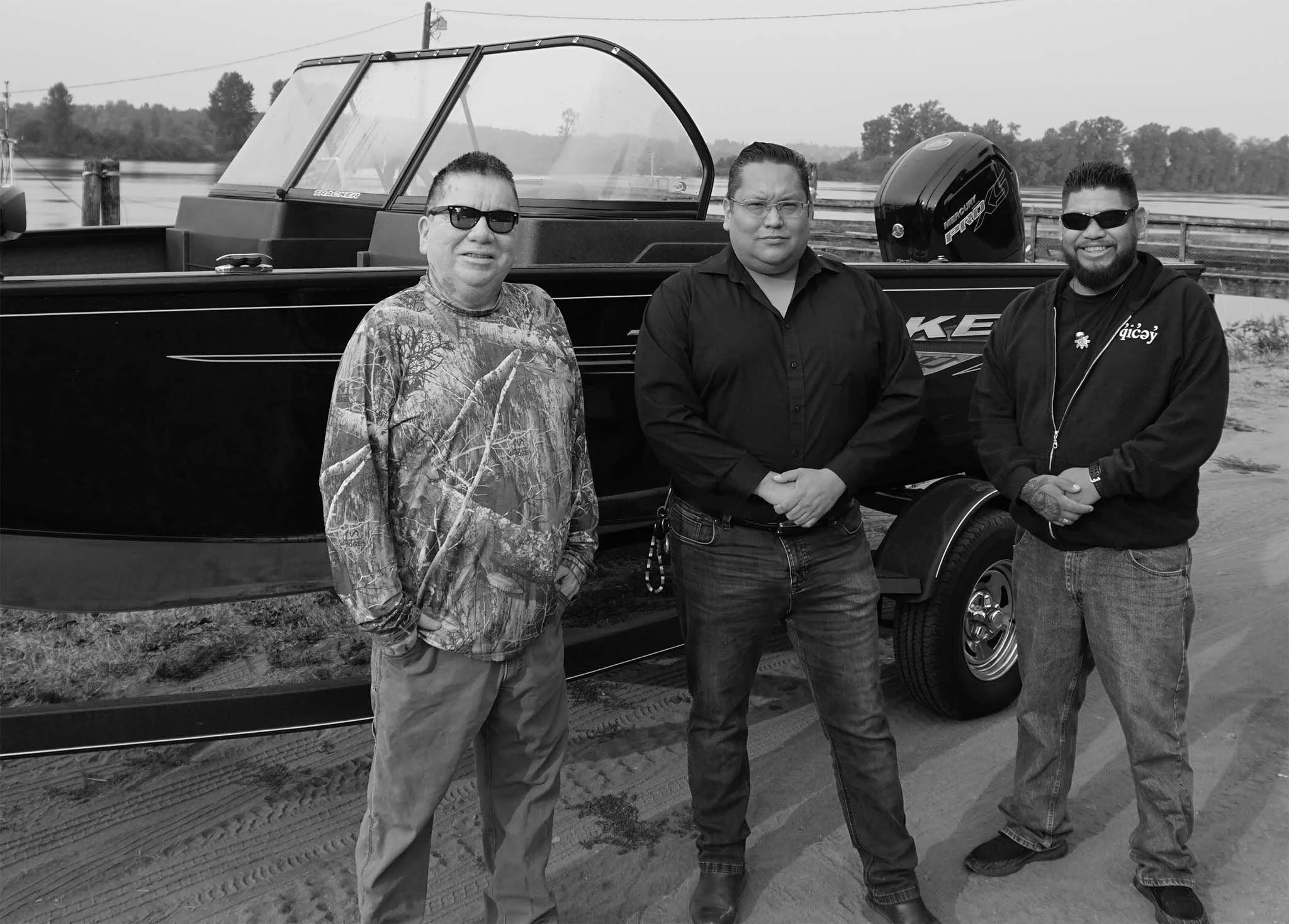 Three men standing outdoors near a boat, smiling and wearing sunglasses, with a water body and trees in the background.