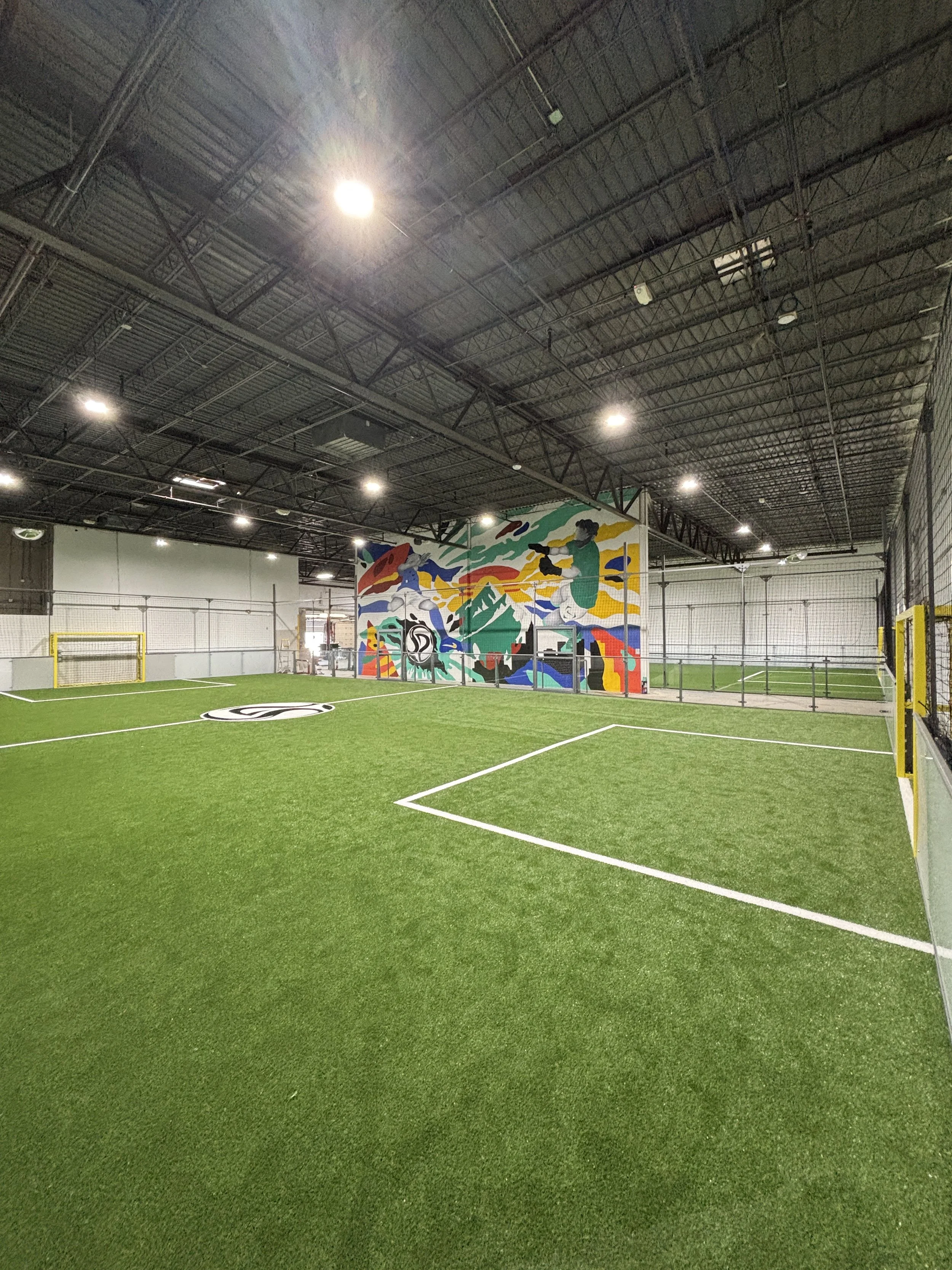 Indoor soccer field with artificial green turf, surrounded by a chain-link fence, featuring a colorful mural wall with abstract shapes and figures, under a high ceiling with industrial lighting.