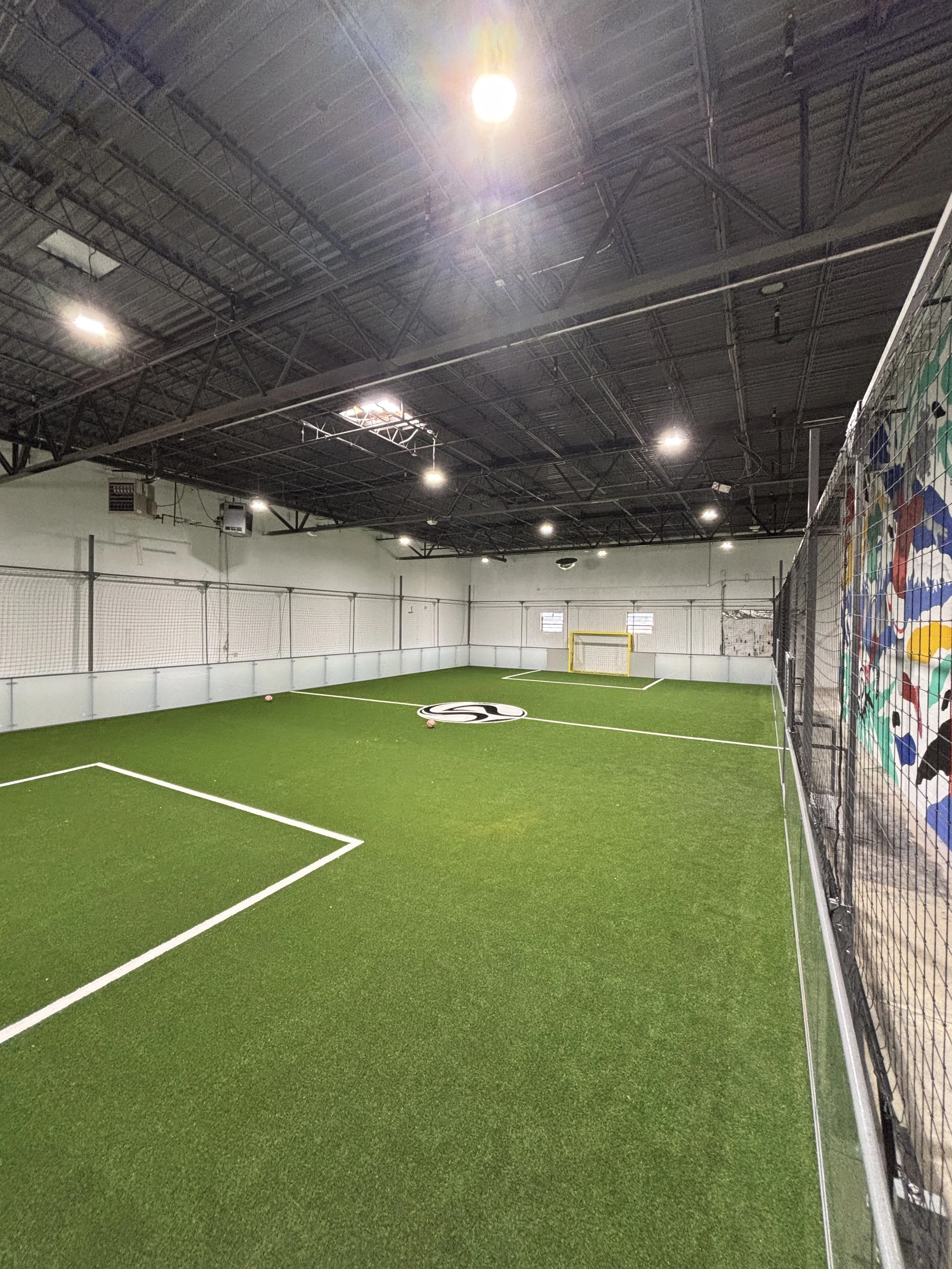 Indoor sports facility with artificial turf field, surrounded by high walls with nets, overhead lighting, and a colorful climbing wall on the right.