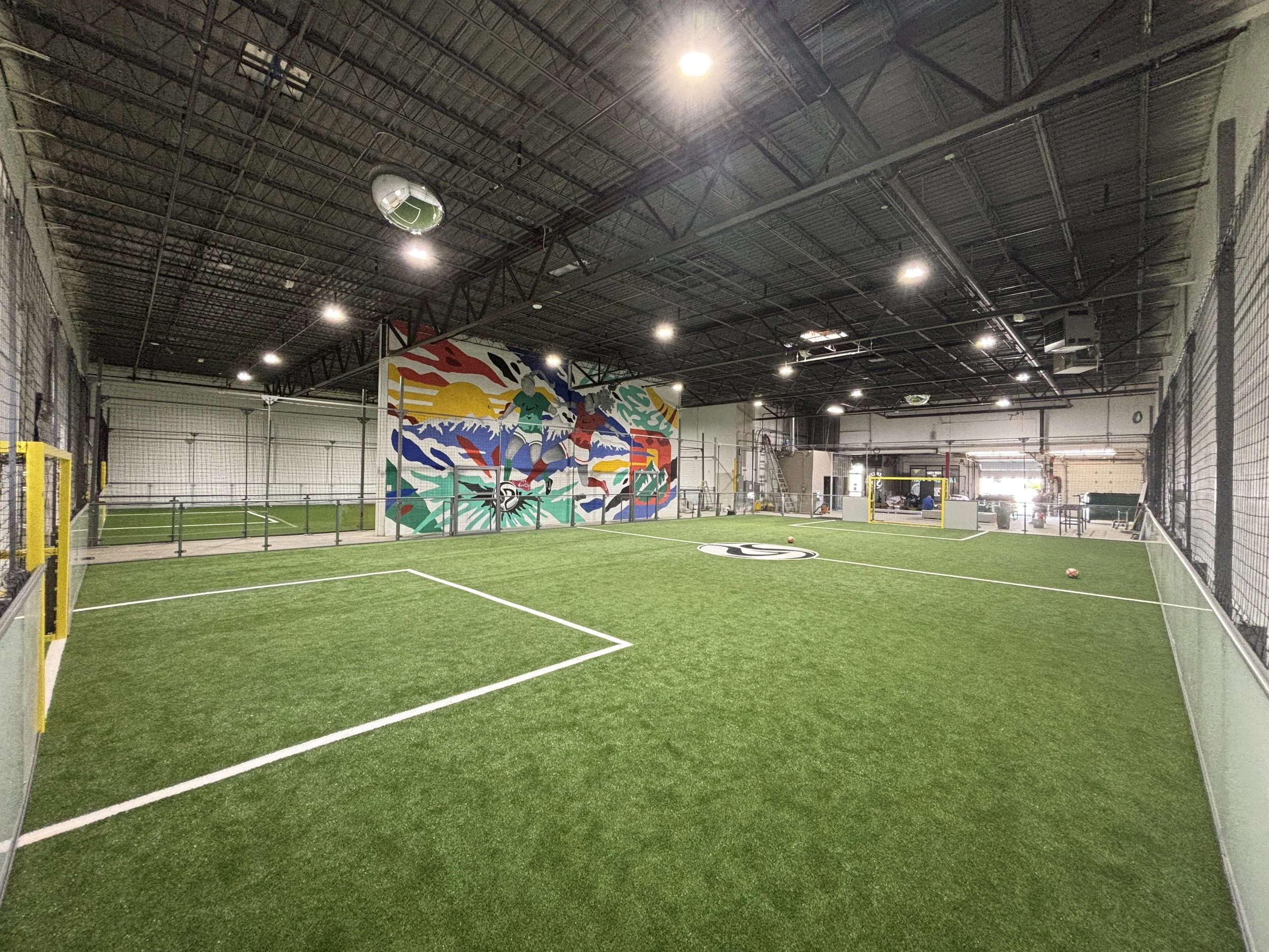 Indoor soccer field with artificial grass, enclosed by black metal fencing and advertising banners, with a small goal at the far end, under a ceiling with netting and bright overhead lighting.