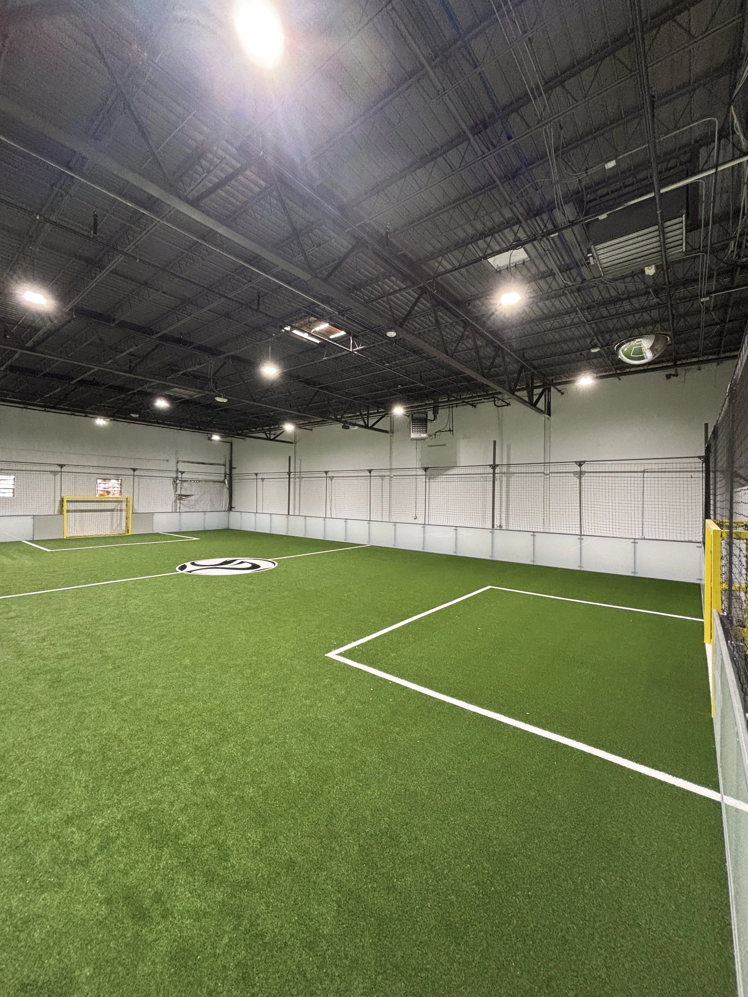 Indoor sports facility with artificial turf, hockey goal net, and white markings on the course. Overhead lighting and visible ceiling structure.