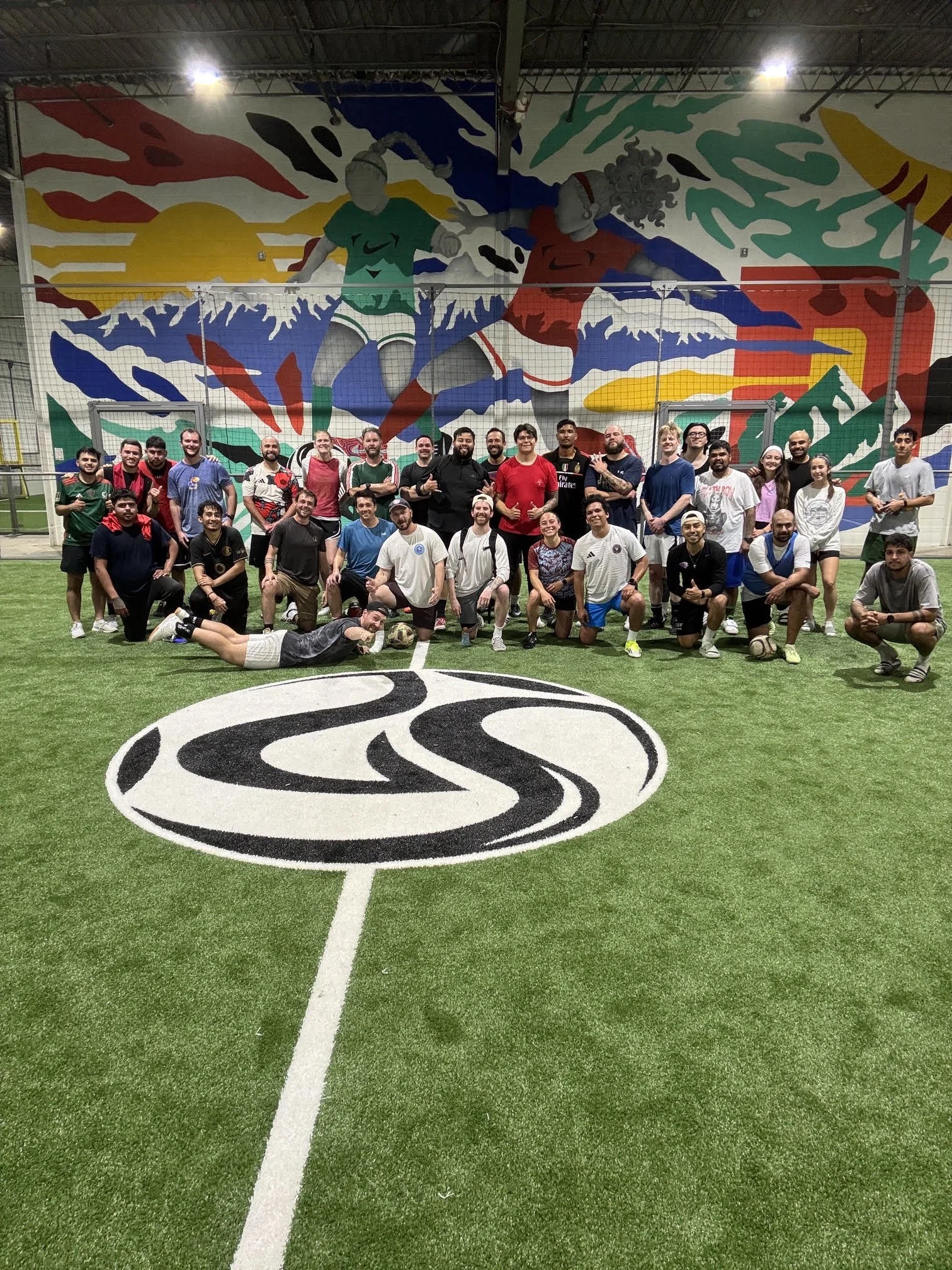 Group of people posing together in an indoor soccer field with a large, colorful mural of two soccer players in the background.