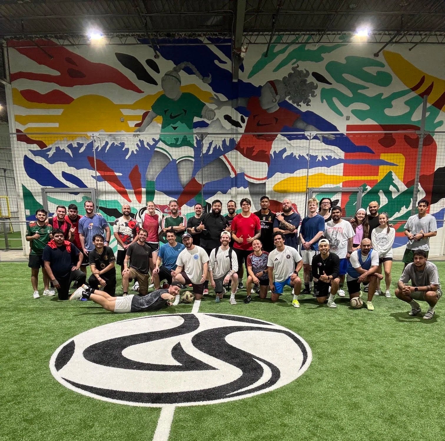 Group of people posing for a photo on an indoor soccer field with a colorful mural of two soccer players jumping for a ball in the background.