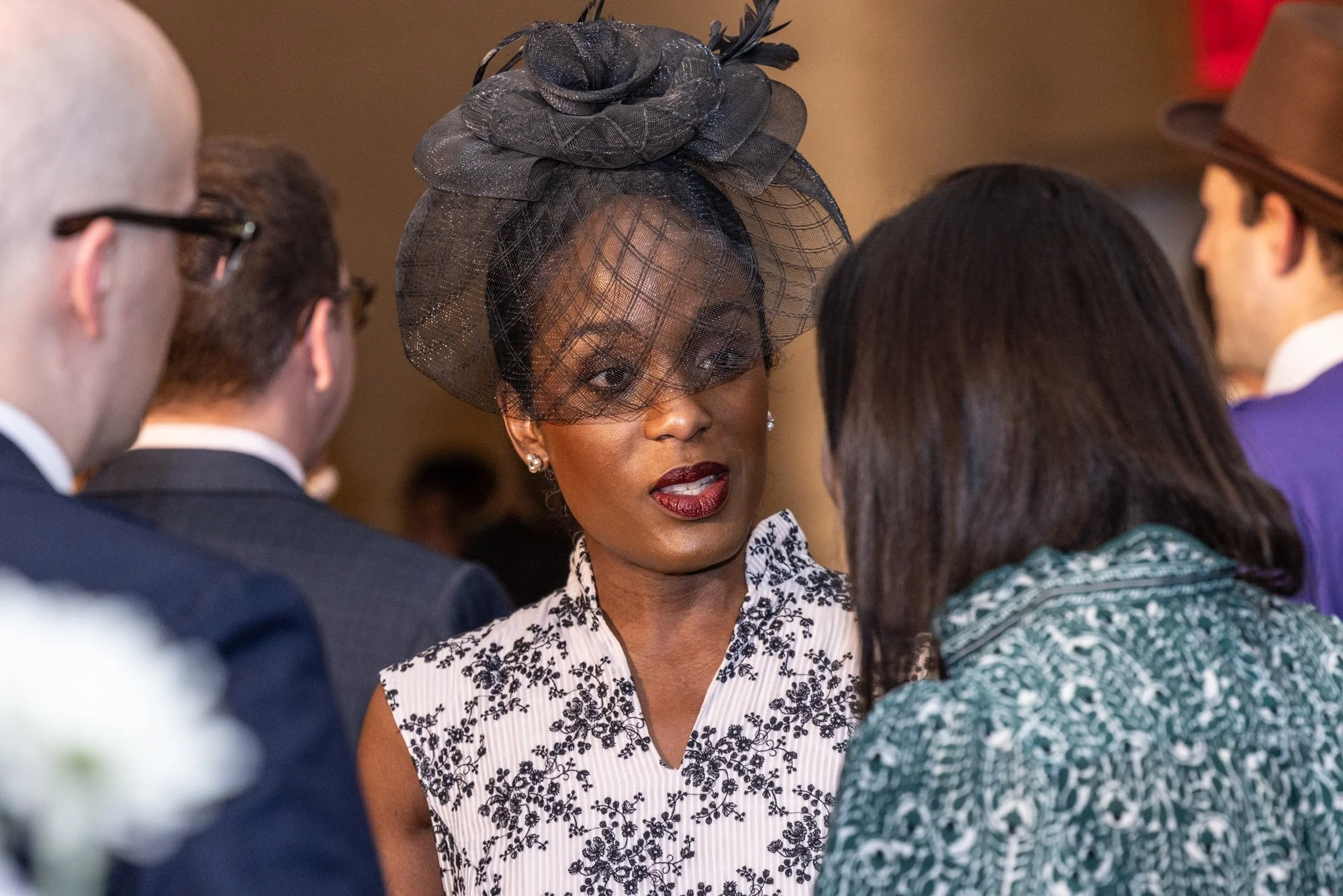 A woman with dark hair and dark lipstick wearing a large black hat with netting, talking to a group of people.