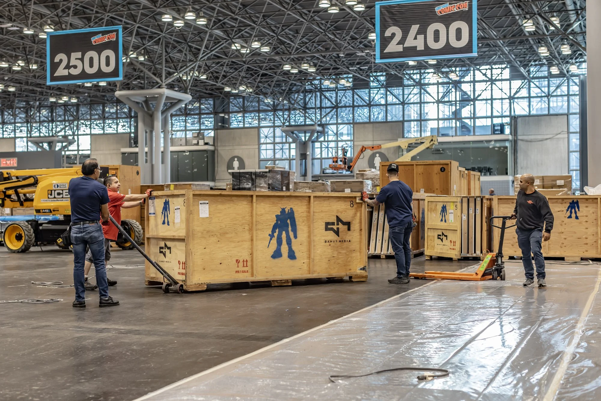 Workers move large wooden shipping crates with pallet jacks inside a convention center, beneath Comic-Con booth signs.