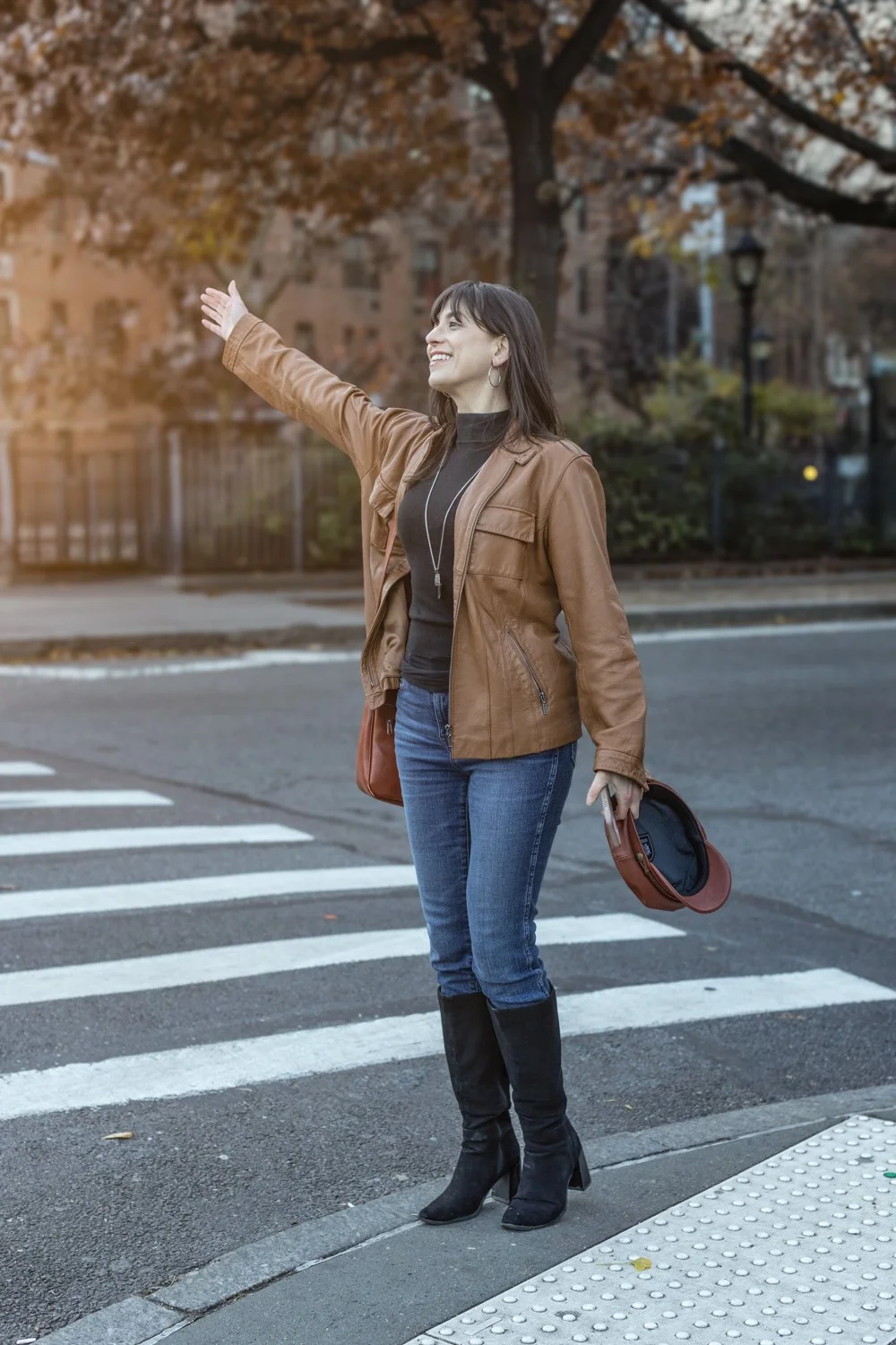 A woman standing at a crosswalk, waving to traffic, in an urban neighborhood during fall, wearing a brown leather jacket, blue jeans, and black knee-high boots.