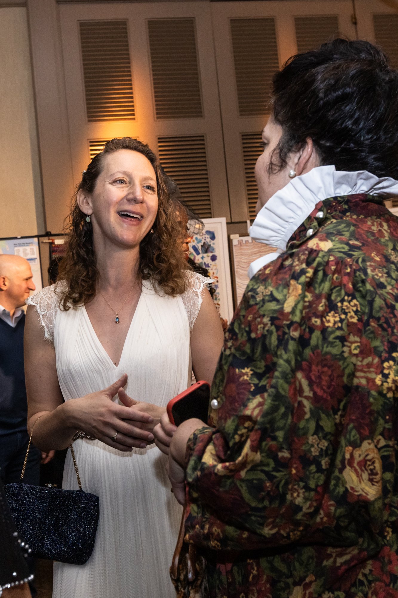 Two women are engaged in a conversation at an indoor event. One woman is dressed in a white dress with lace details and is smiling, while the other woman, dressed in a floral jacket with pearl earrings, is holding a smartphone. In the background, the
