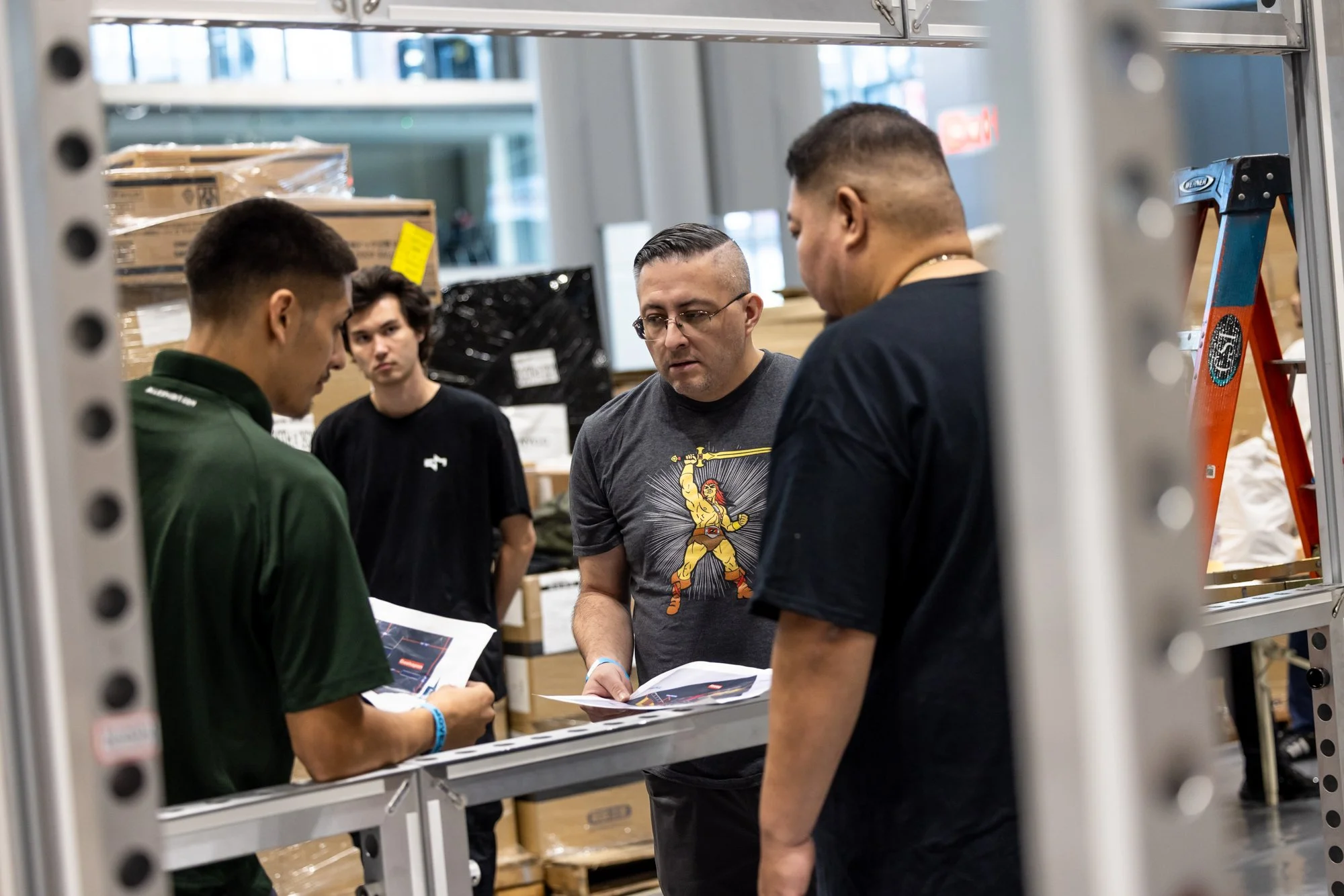 Four men review papers in a warehouse surrounded by cardboard boxes and a step ladder.