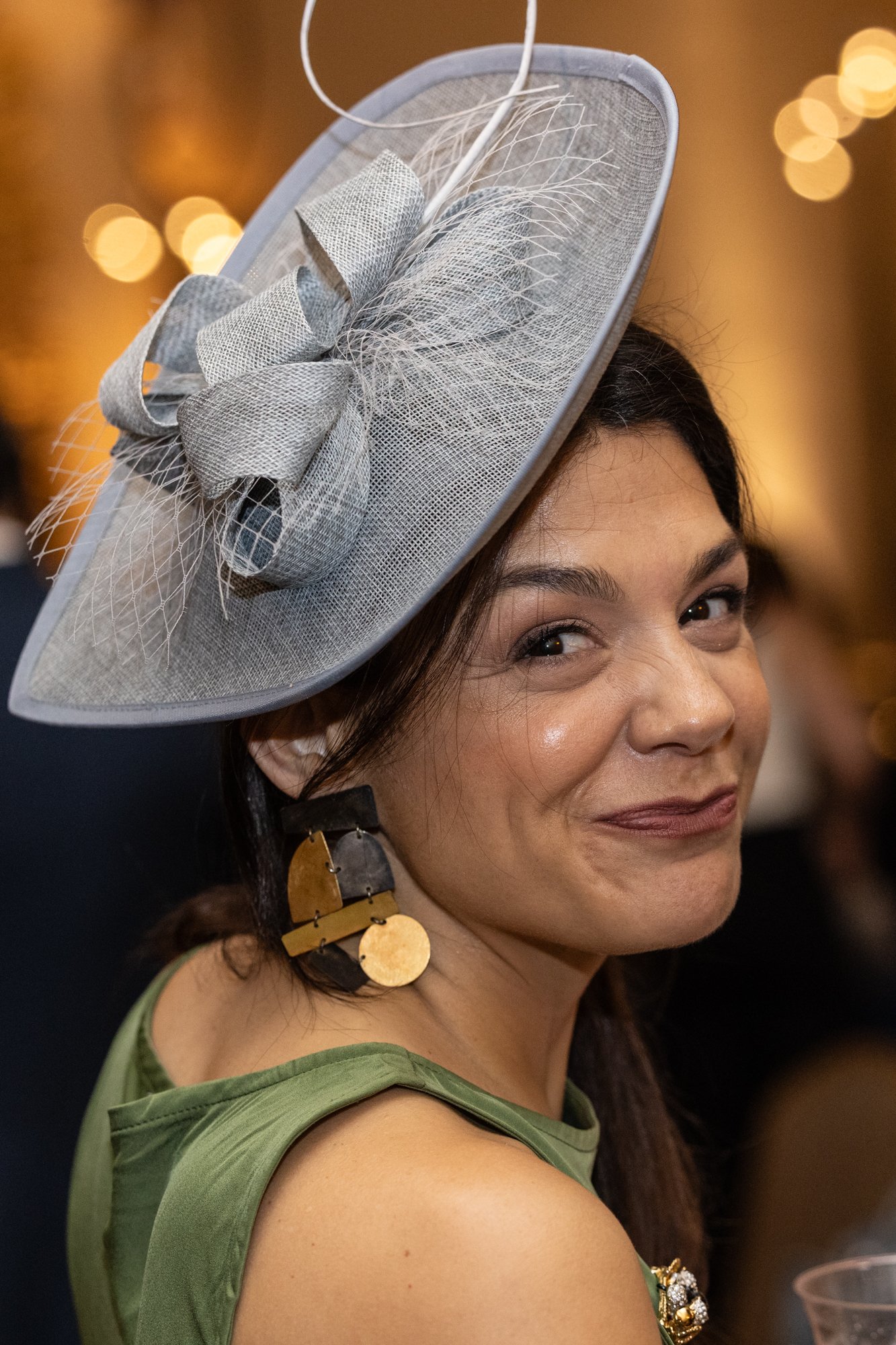 A woman with dark hair and a light skin tone smiling, wearing a large gray hat with a bow and feather embellishments, gold and black earrings, and a green sleeveless top.