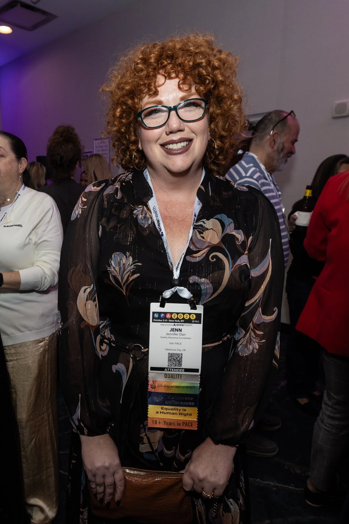 A woman with curly red hair and glasses smiling at a conference or event, wearing a badge with her name Jennifer Dan and standing in front of a group of people.