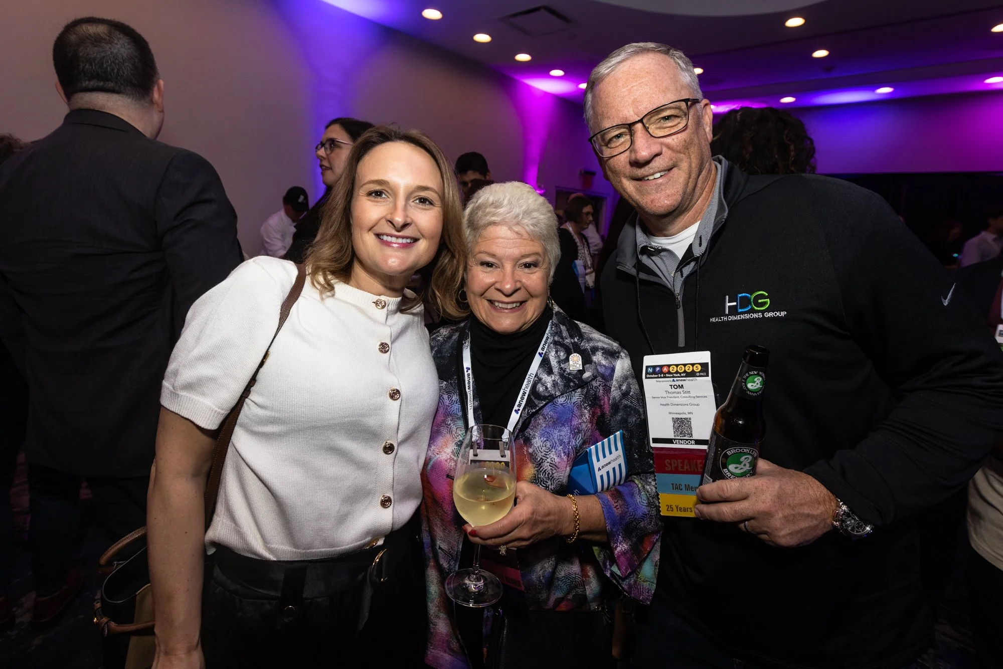 Three people smiling at a social event, with a woman in a white top, an older woman holding a glass of white wine, and a man wearing glasses and a black jacket with a conference badge, in a dimly lit room with purple lighting.