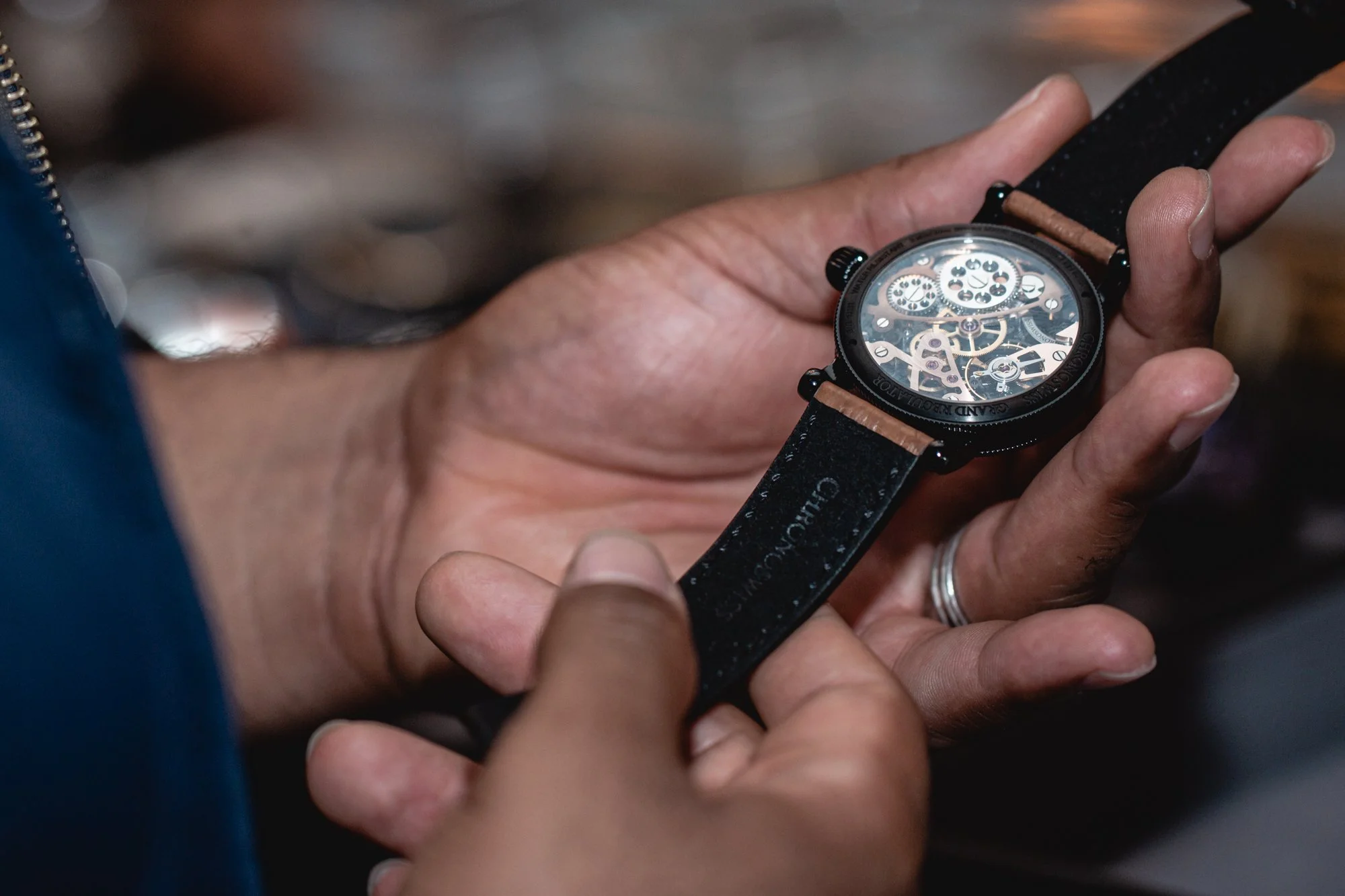 Close-up of a person holding a skeleton watch with visible gears and mechanical parts, wearing a silver ring on index finger, with a blurred background.