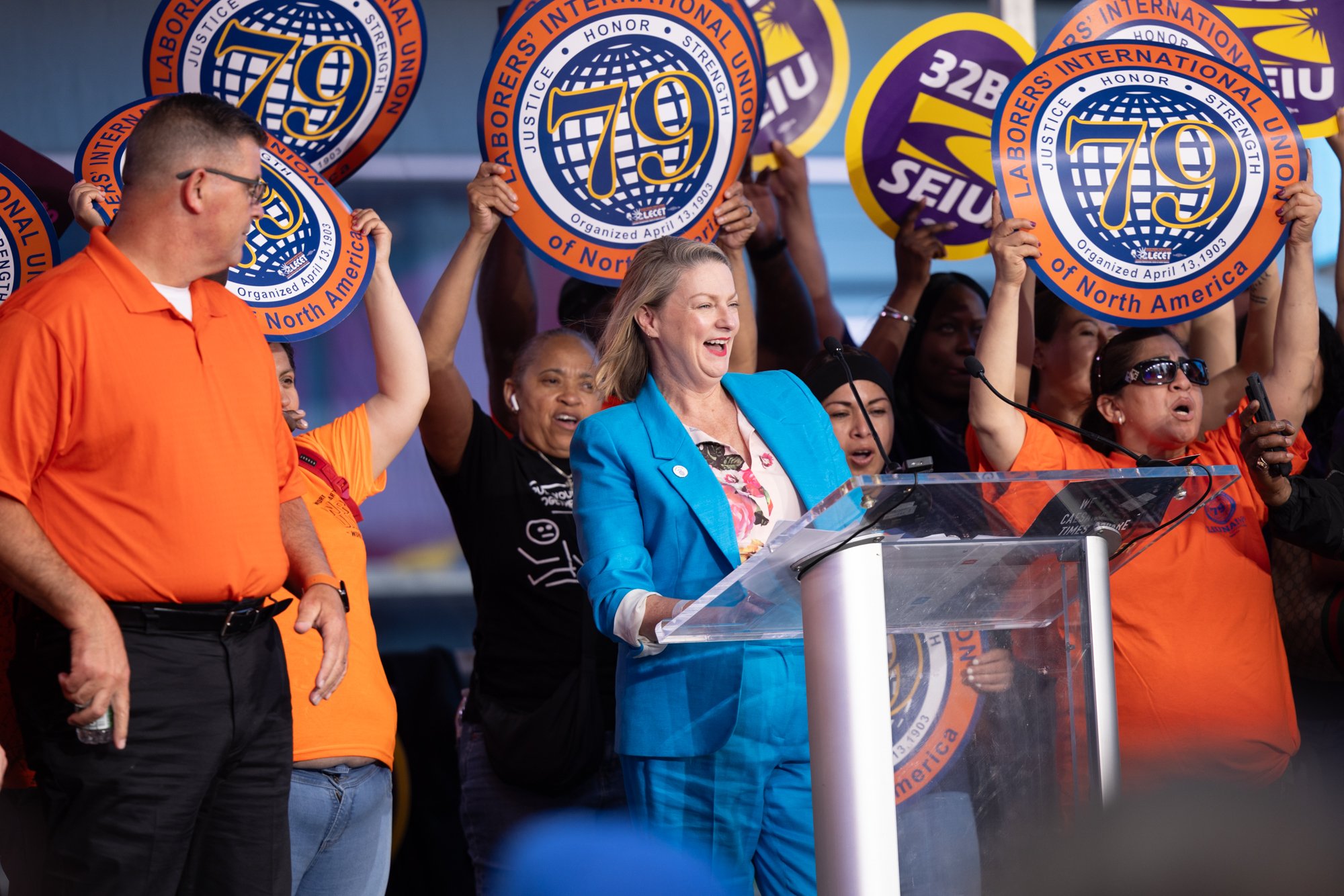 A group of people, including a woman at a podium, holding signs with the number 79 and logos for Laborers' International Union of North America, during a rally or event.