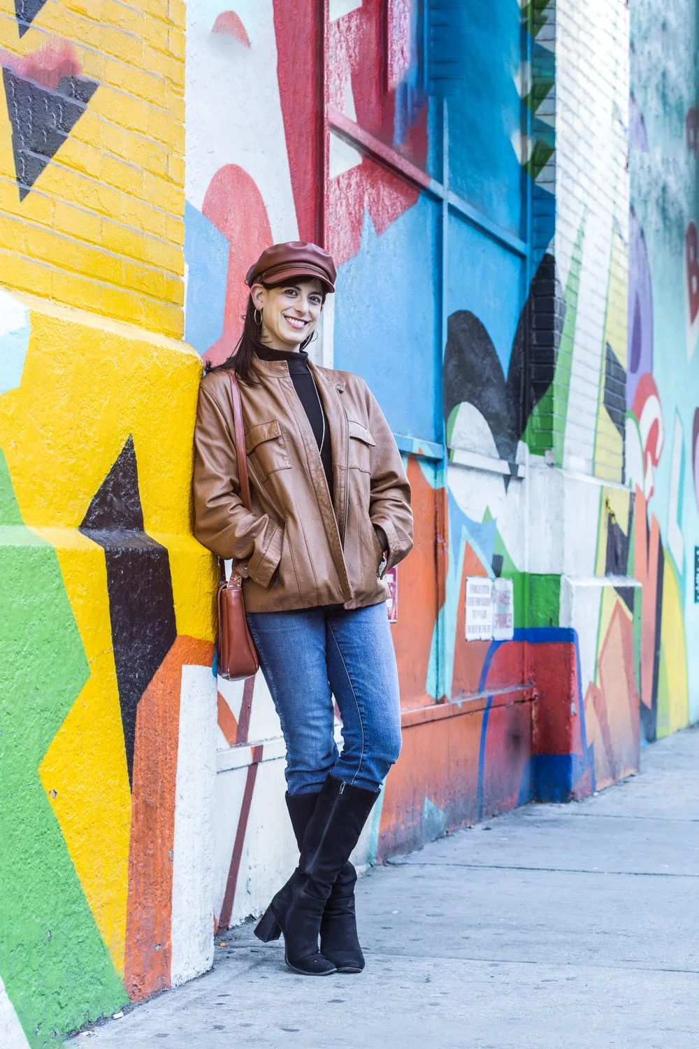 A woman in a brown leather jacket and black boots leaning against a colorful mural wall on city street