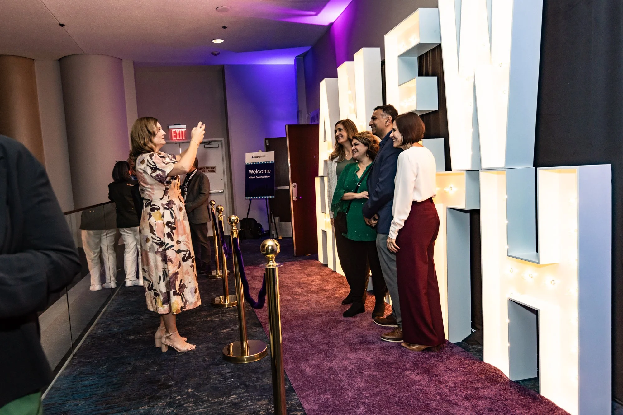 A woman in a floral dress taking a photo of four smiling people standing in front of a large illuminated sign at an event.