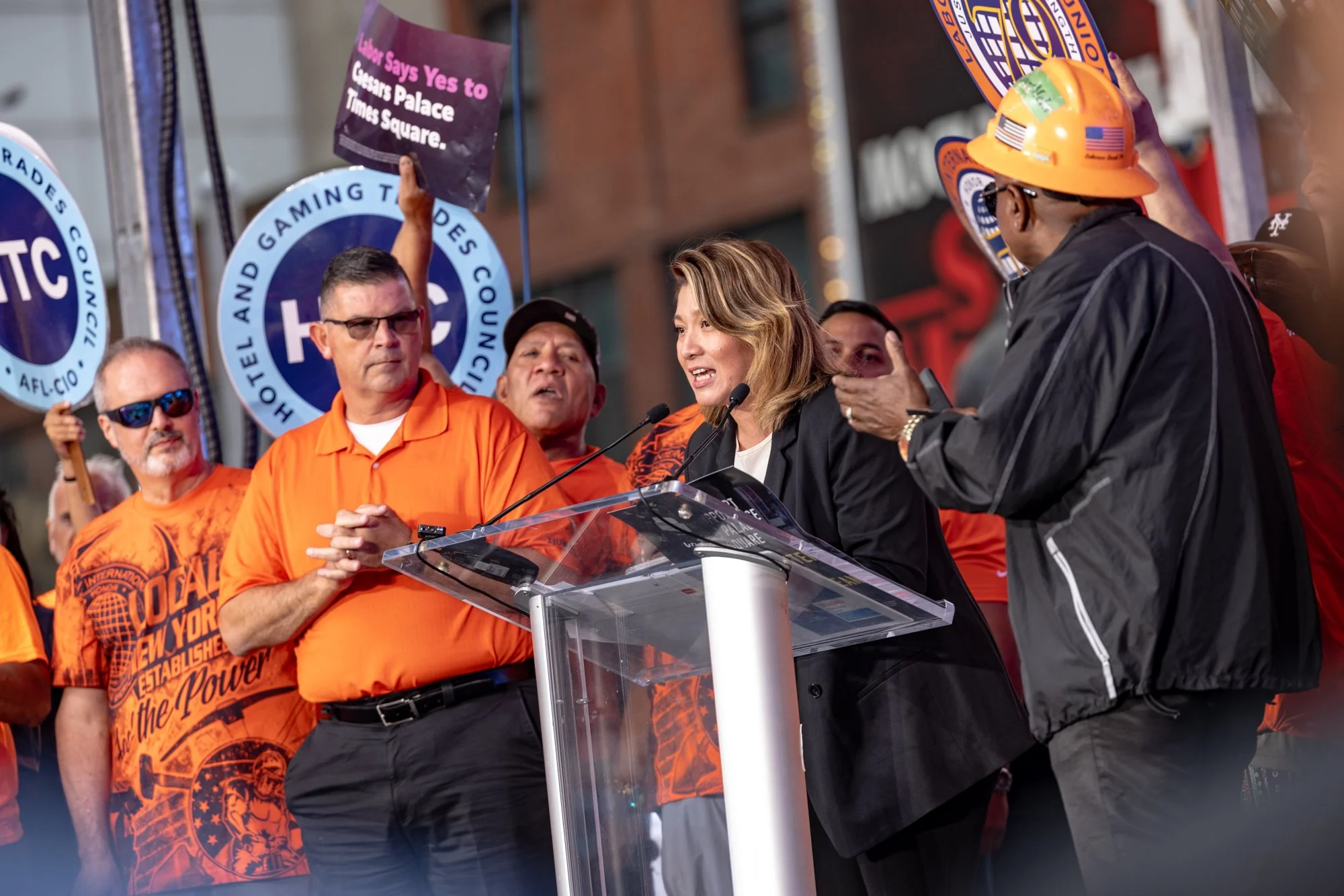 A woman speaking at a podium during a public event, surrounded by a group of people including protestors holding signs, with some people wearing orange shirts and a person wearing a yellow hard hat.