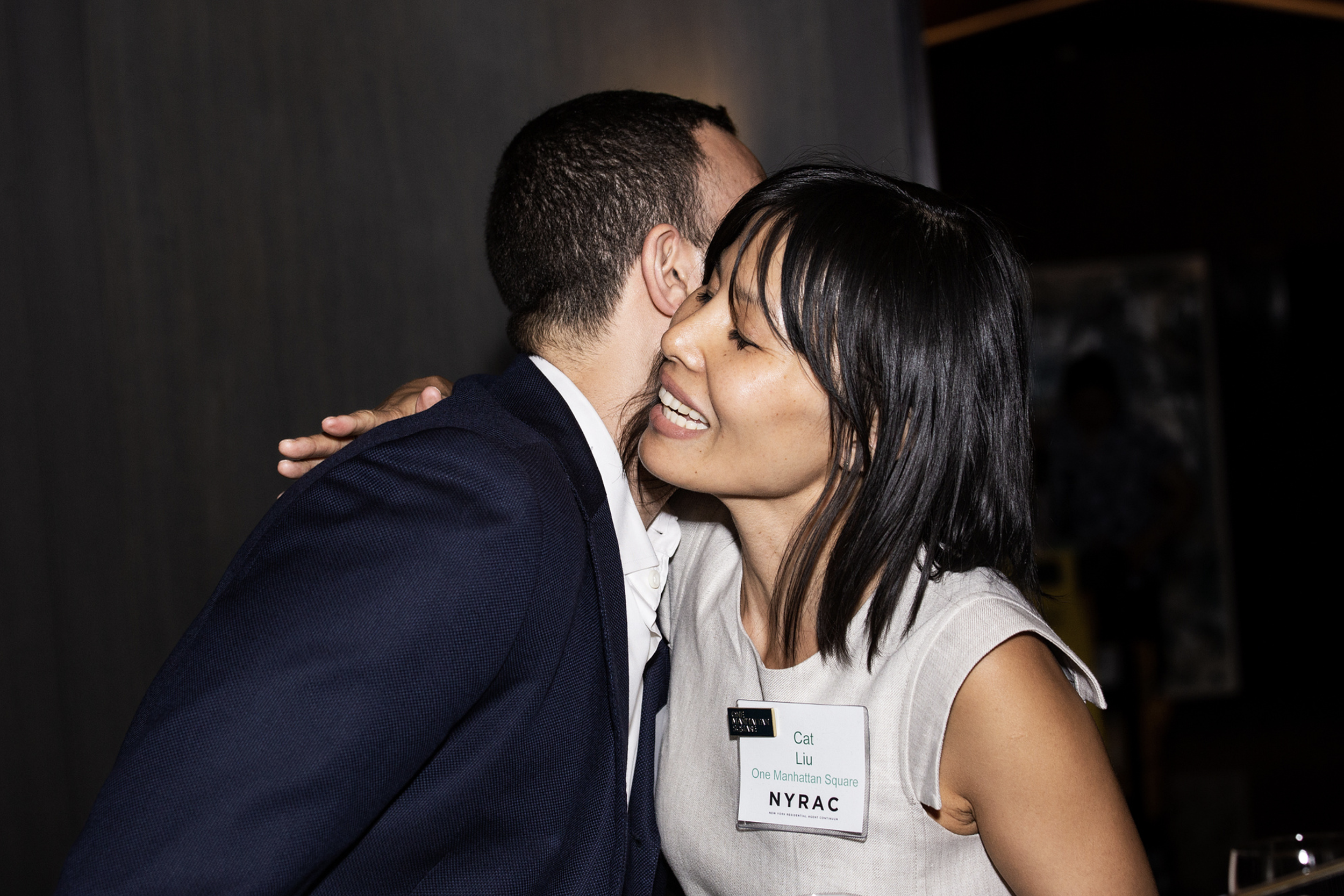 A man and a woman hugging each other, smiling with eyes closed, at an indoor event. The woman has a name tag reading 'Cat Liu' and is wearing a light-colored dress.