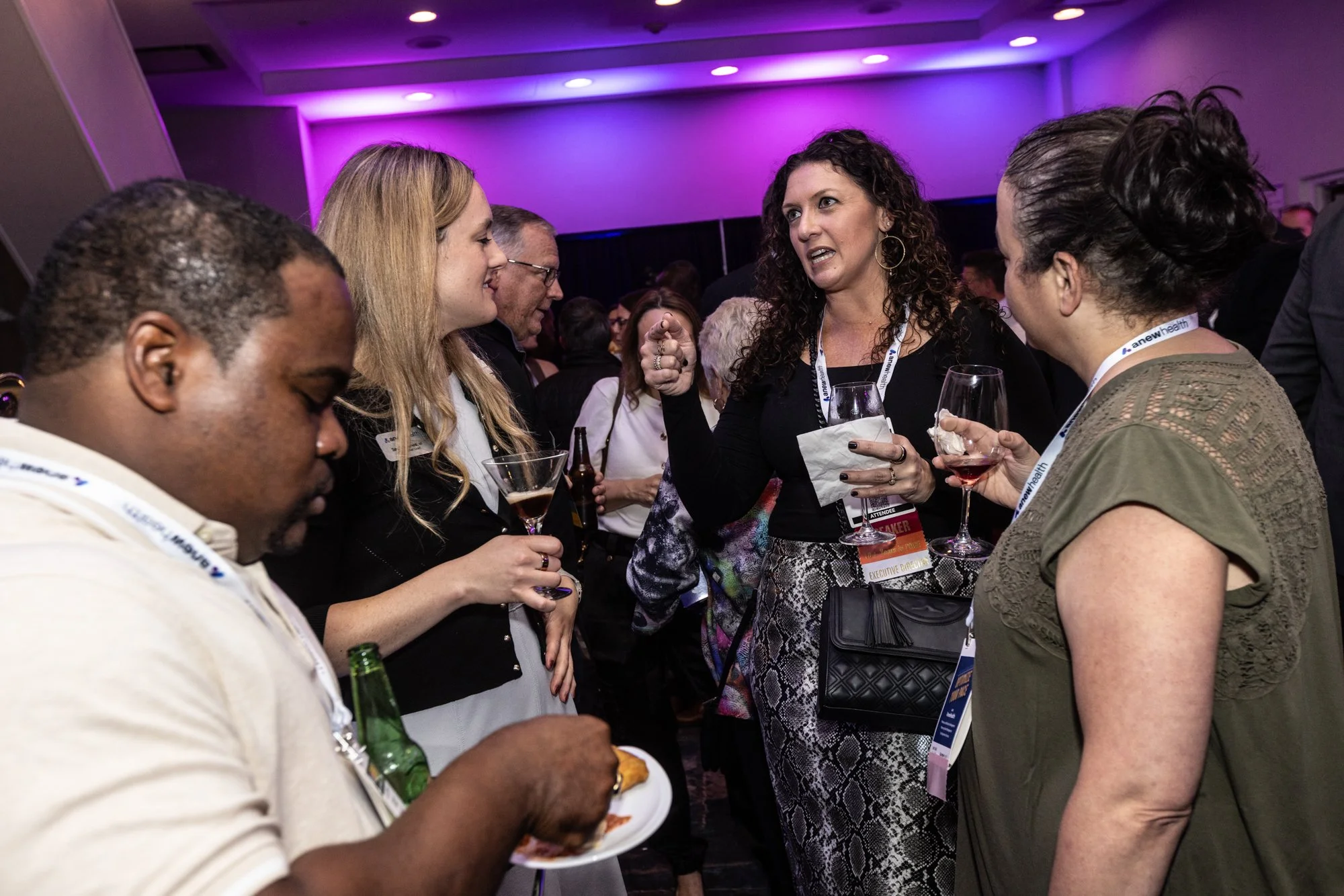 A group of people at a social event, engaging in conversation. They are holding drinks and wearing conference badges.